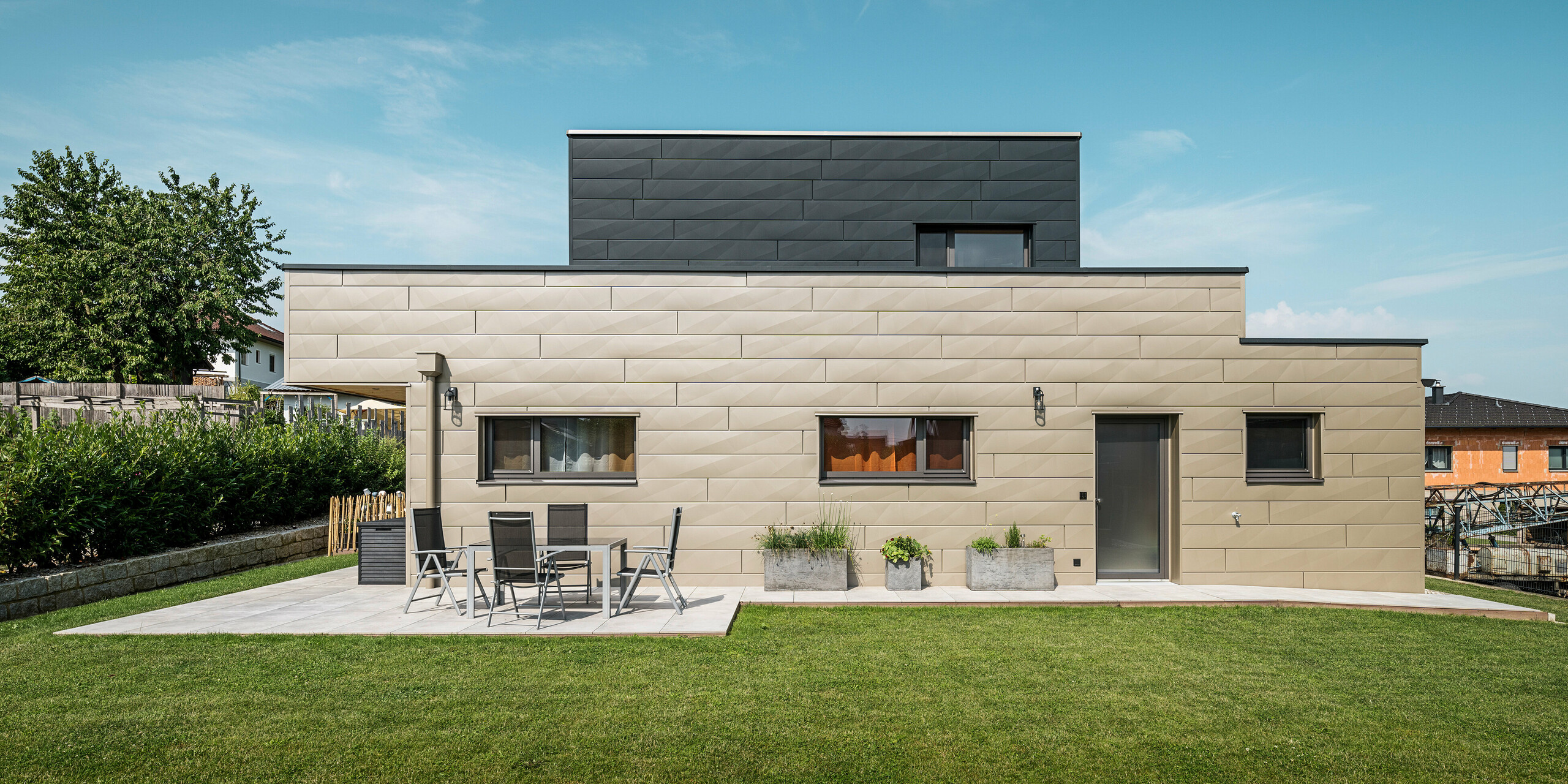 Vue sur le jardin d'une maison familiale moderne à Wippenham, en Autriche, avec une façade en aluminium PREFA bicolore. La façade est constituée de panneaux de façade Siding.X dans les couleurs bronze et anthracite, qui forment un contraste élégant et confèrent au bâtiment des lignes modernes et claires. Au premier plan se trouve une terrasse spacieuse avec mobilier de jardin qui complète la pelouse bien entretenue et la conception architecturale moderne de la maison.