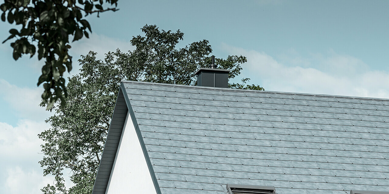 Le toit à deux pans de la maison individuelle classique a été recouvert avec la nouvelle tuile R.16 PREFA couleur gris pierre. Trois fenêtres de toit ont été intégrées dans la surface de toit et des arrêts neige ont été montés. La façade a été simplement laissée blanche.