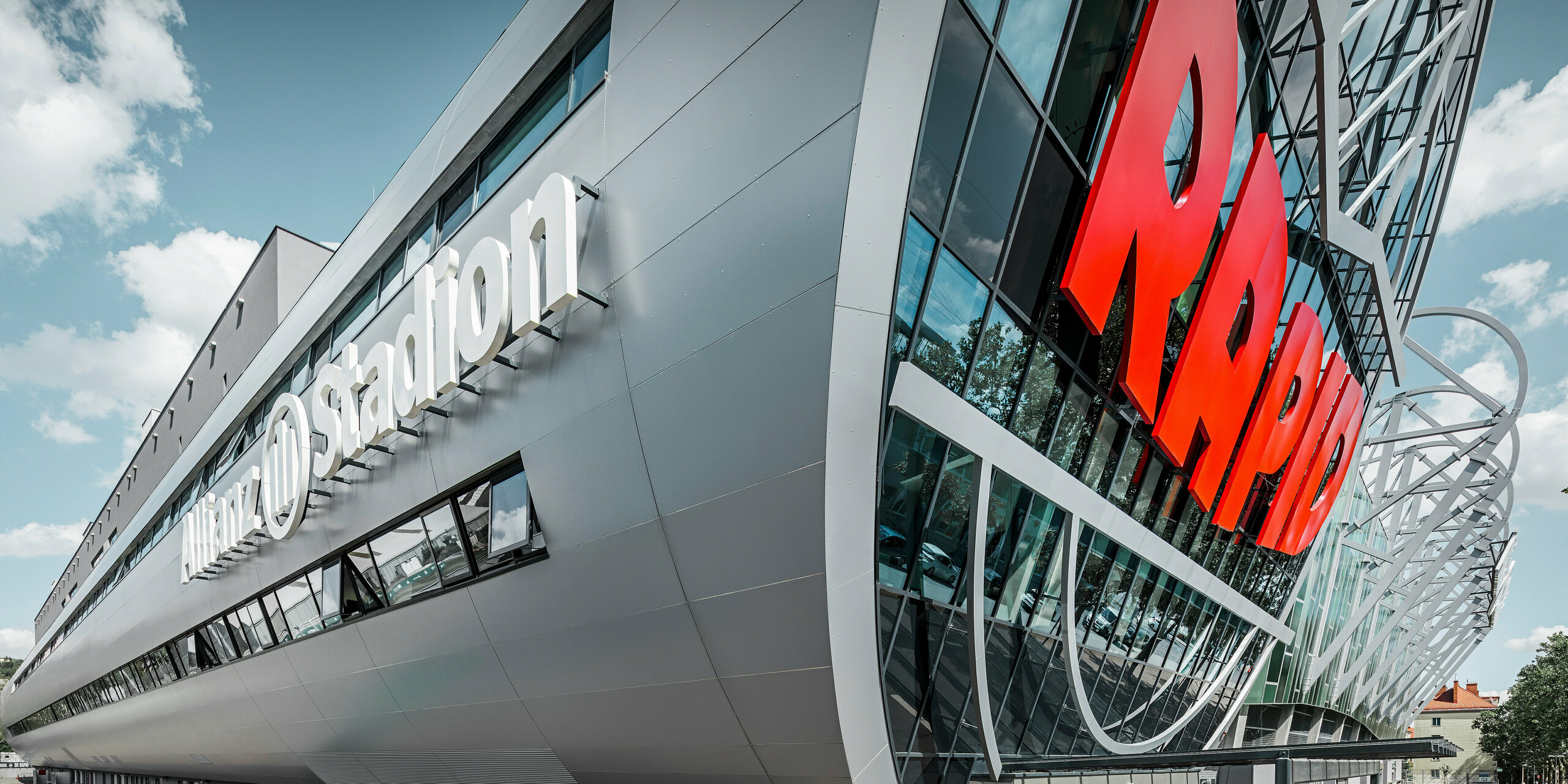 Nahaufnahme der Fassade des Allianz Stadions, Heimat des SK Rapid Wien. Die eindrucksvolle Röhre im Eingangsbereich präsentiert die PREFA Aluminium Verbundplatten in Silbermetallic. Der Blickwinkel unterstreicht die moderne Architektur. Durch den strahlend roten RAPID-Schriftzug und die klare Linienführung der silbernen Platten erhält die Röhre eine visuell beeindruckende Dynamik und Ästhetik. Auf diese Weise wird bereits beim Gang zum Stadion bei allen Fußball-Fans Leidenschaft entfacht.