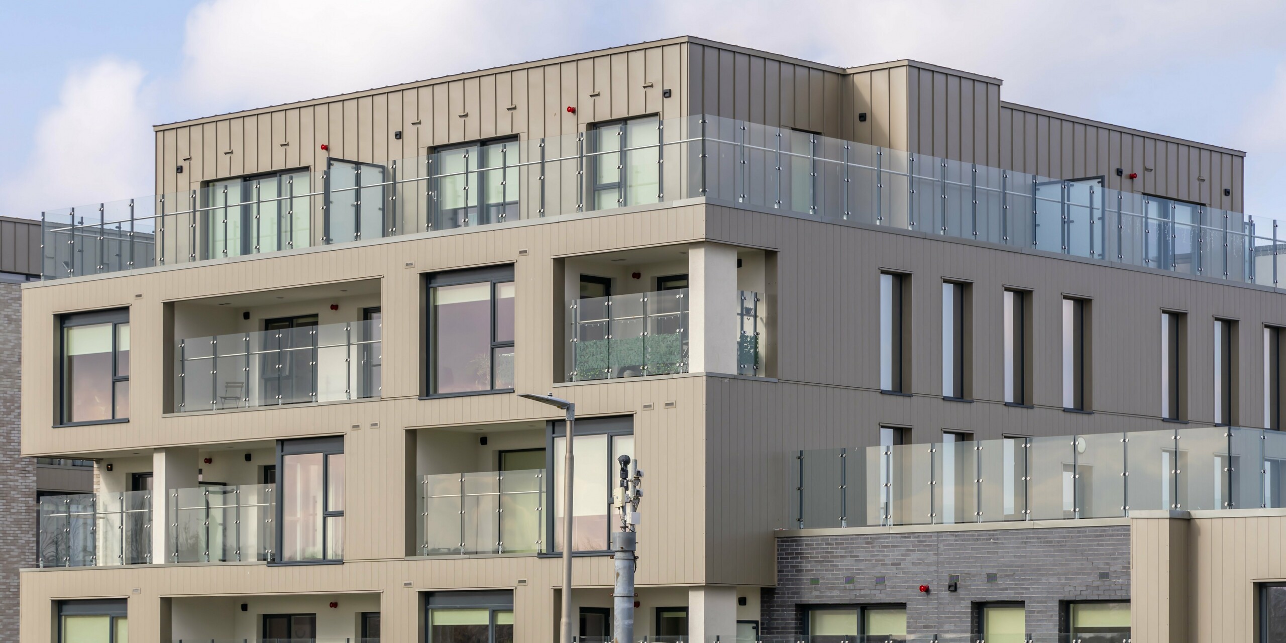 Complexe résidentiel Palmers Gate à Palmerstown, Dublin, Irlande - gros plan d'un immeuble résidentiel avec parements PREFA de couleur bronze et système de façade PREFALZ. Les balcons sont équipés d'élégantes balustrades en verre qui soulignent le confort de vie moderne. La façade combine un revêtement en aluminium de couleur bronze avec des éléments en brique, créant un aspect harmonieux et attrayant. Le complexe immobilier offre aux résidents des espaces extérieurs de haute qualité et impressionne par son architecture robuste et esthétique.