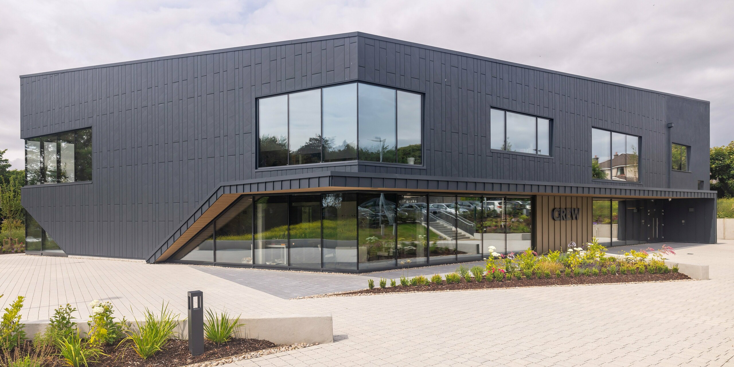 Vue frontale du bâtiment CREW à Galway, en Irlande, avec une façade moderne en aluminium PREFA en Siding.X et PREFALZ en anthracite P.10. Les lignes inclinées et les grandes fenêtres caractérisent la conception architecturale sophistiquée du bâtiment. Le bardage en aluminium anthracite combiné confère au bâtiment une esthétique élégante et contemporaine, complétée par un aménagement paysager bien entretenu.