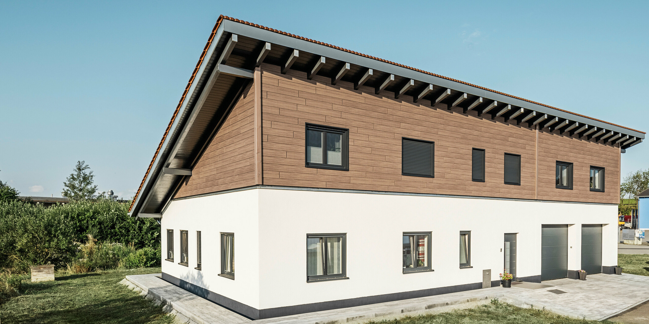 Schräger Blick auf ein Einfamilienhaus mit einer Aluminiumfassade in einer Holzoptik am Obergeschoss. Das untere Stockwerk des Gebäudes mit Pultdach ist in schlichtem Weiß gehalten. 