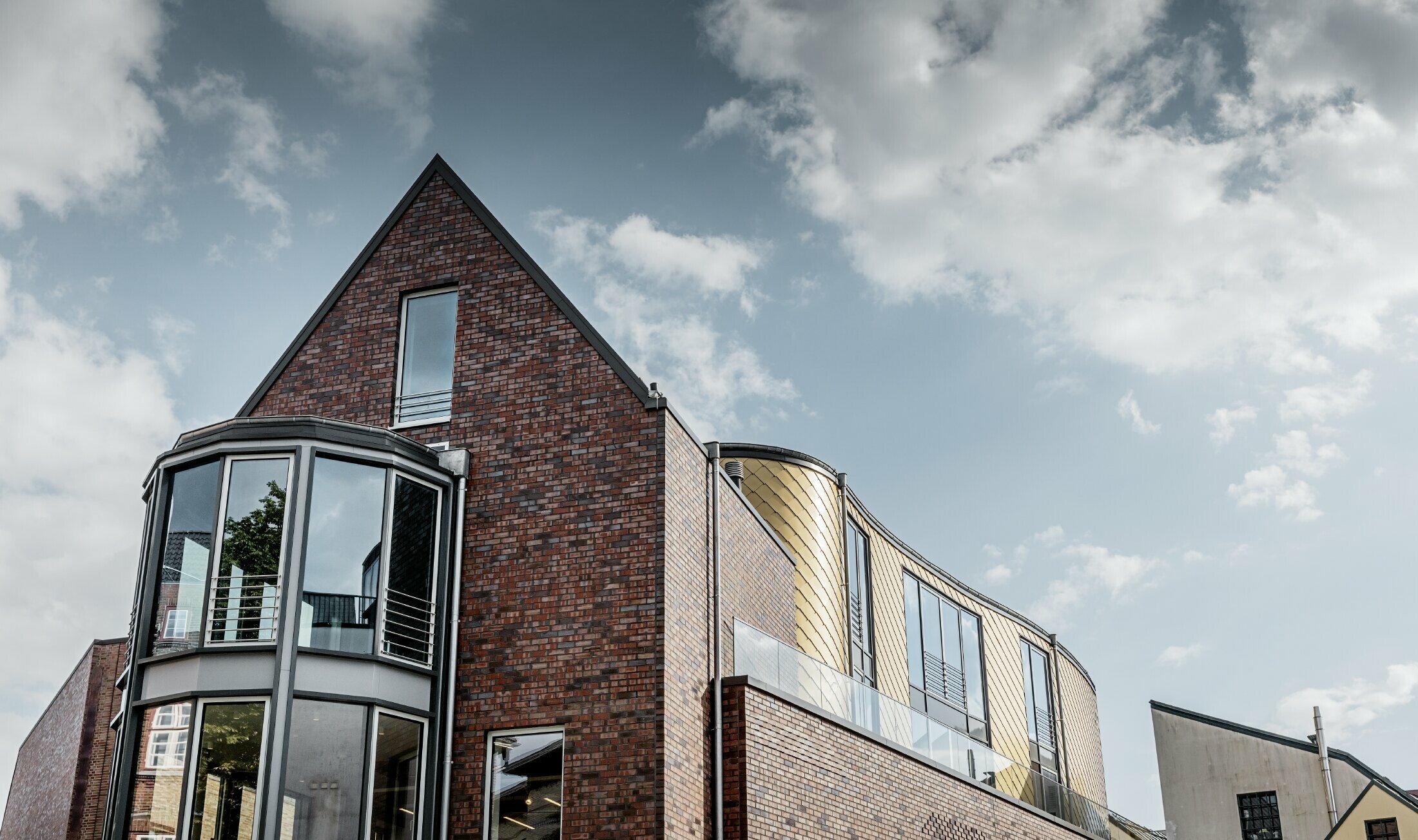 Zakenpand met baksteengevel en royale glasoppervlakken; Een deel van de gebogen gevel is bekleed met de goudkleurige aluminium gevellosange van PREFA.