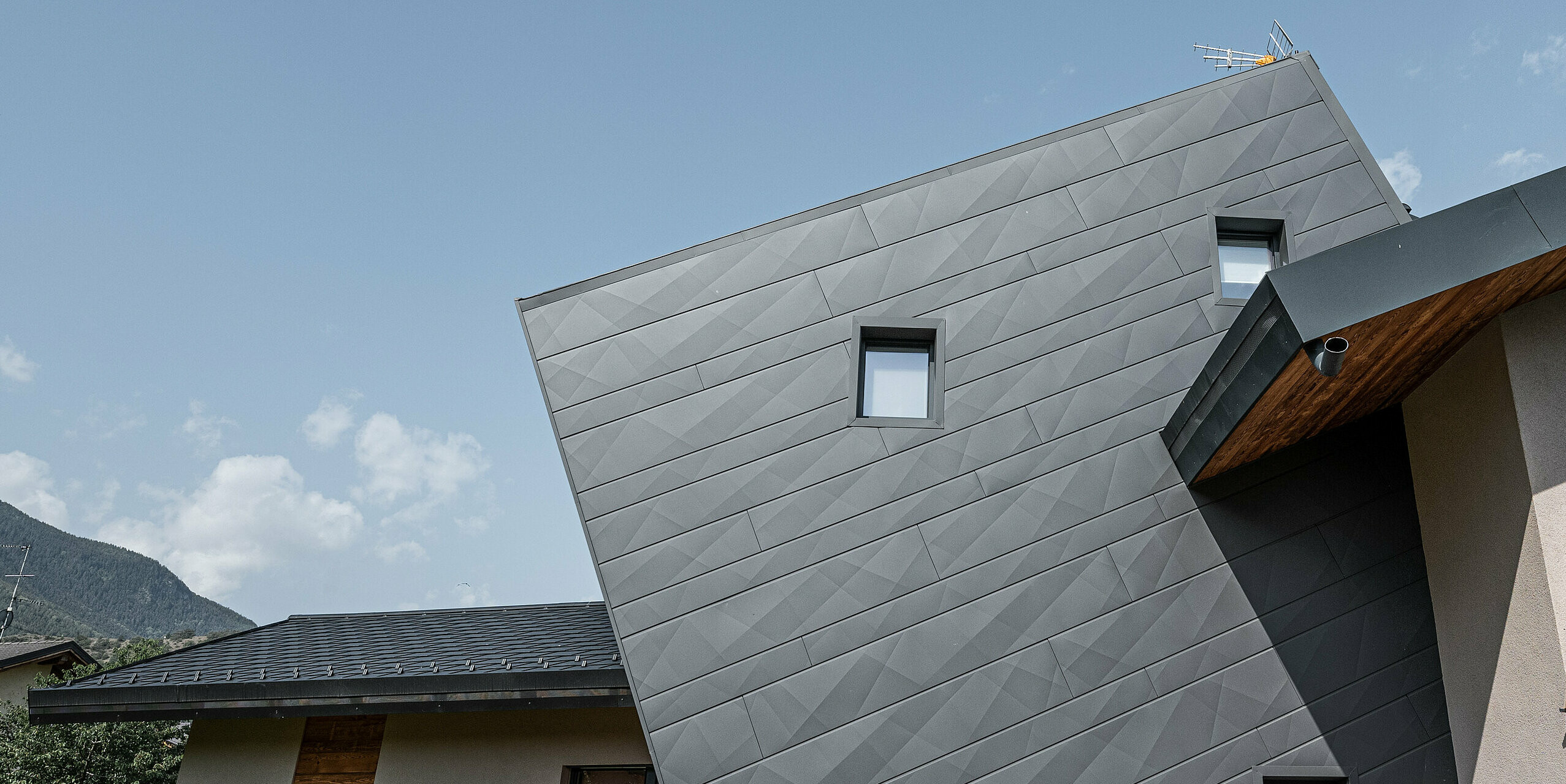 Die Villa Tia in Gressan, Italien, zeigt eine moderne Fassadengestaltung mit PREFA Siding.X in P.10 Dunkelgrau. Die dynamische Anordnung der Paneele erzeugt ein dreidimensionales Erscheinungsbild, das durch den Kontrast zu den natürlichen Holzelementen und der umgebenden Landschaft hervorgehoben wird. Diese innovative Sonderlösung der schrägen Siding.X Fassadenpaneele steht für ein zeitgemäßes und langlebiges Design.
