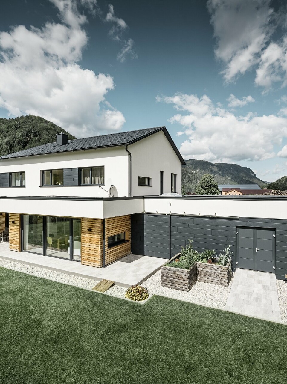 Das Bild zeigt ein modernes Einfamilienhaus in einer idyllischen Landschaft mit einer klaren Sicht auf bewaldete Hügel im Hintergrund. Das Haus zeichnet sich durch eine Kombination aus weißen Putzwänden und einer markanten Fassadenverkleidung mit PREFA Siding.X in P.10 Anthrazit aus, die für einen modernen und langlebigen Look sorgt. Die dunkle Färbung der Fassadenelemente bildet einen ansprechenden Kontrast zum hellen Außendesign und zur natürlichen Umgebung. Die Anordnung der Siding.X Fassadenpaneele ist präzise und verleiht der Immobilie einen strukturierten und hochwertigen Charakter, der typisch für PREFA Produkte ist. Das Dach des Gebäudes wurde mit der bewährten PREFA Dachplatte eingedeckt. Die Dachentwässerung erfolgt durch das Ablaufrohr und die Hängerinne von PREFA.