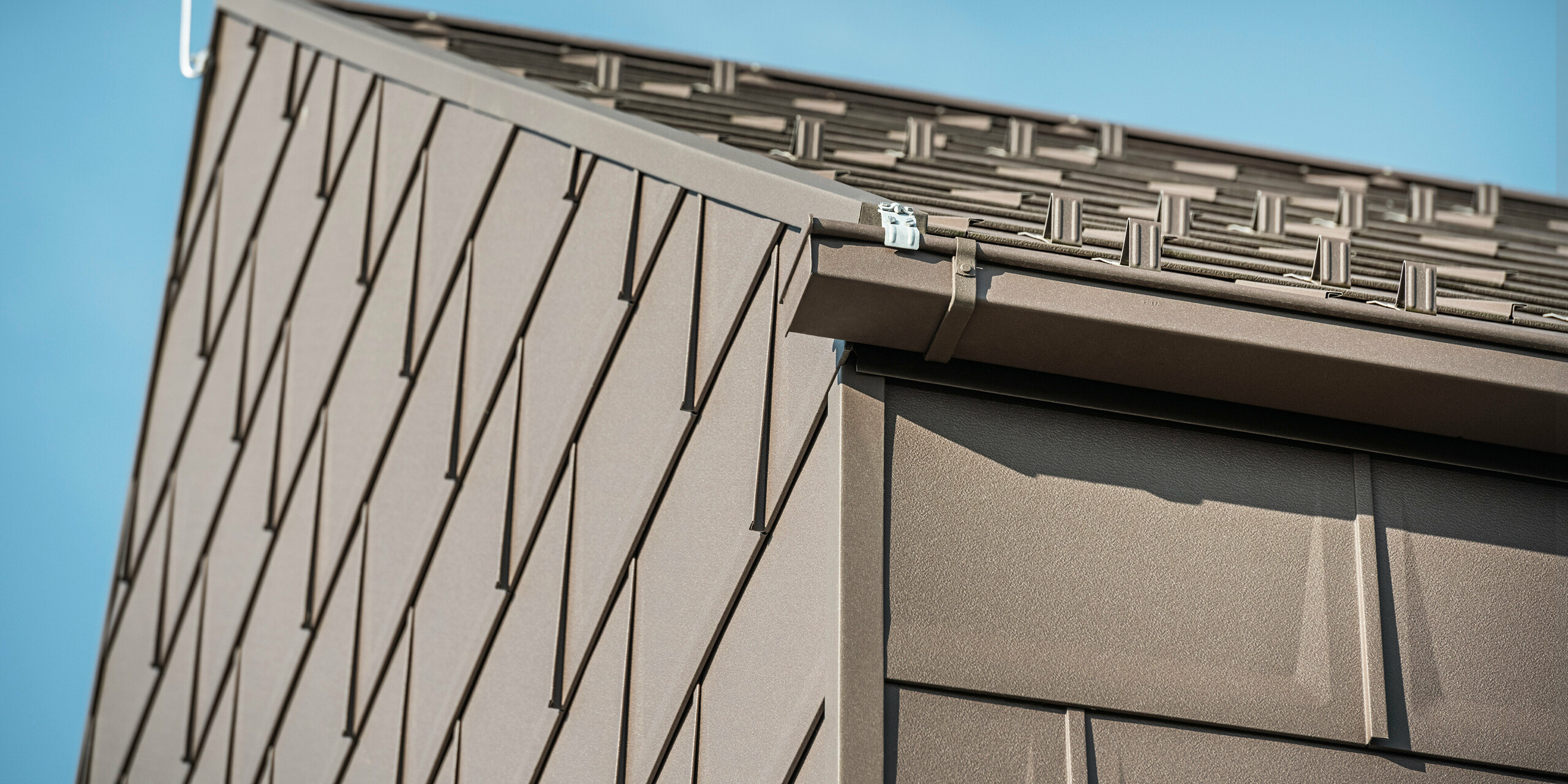 Detailaufnahme der Dachentwässerung eines Hauses in Neukirchen, Österreich, mit einer PREFA Kastenrinne. Das Dachpaneel R.16 in der braunen Farbe P.10 bildet nicht nur das Dach, sondern schmückt auch die Fassade und schafft ein einheitliches und elegantes Erscheinungsbild. Diese Nahaufnahme zeigt die Präzision und Qualität der PREFA Aluminiumprodukte, die Design und Funktionalität für ein modernes Wohnkonzept verbinden.