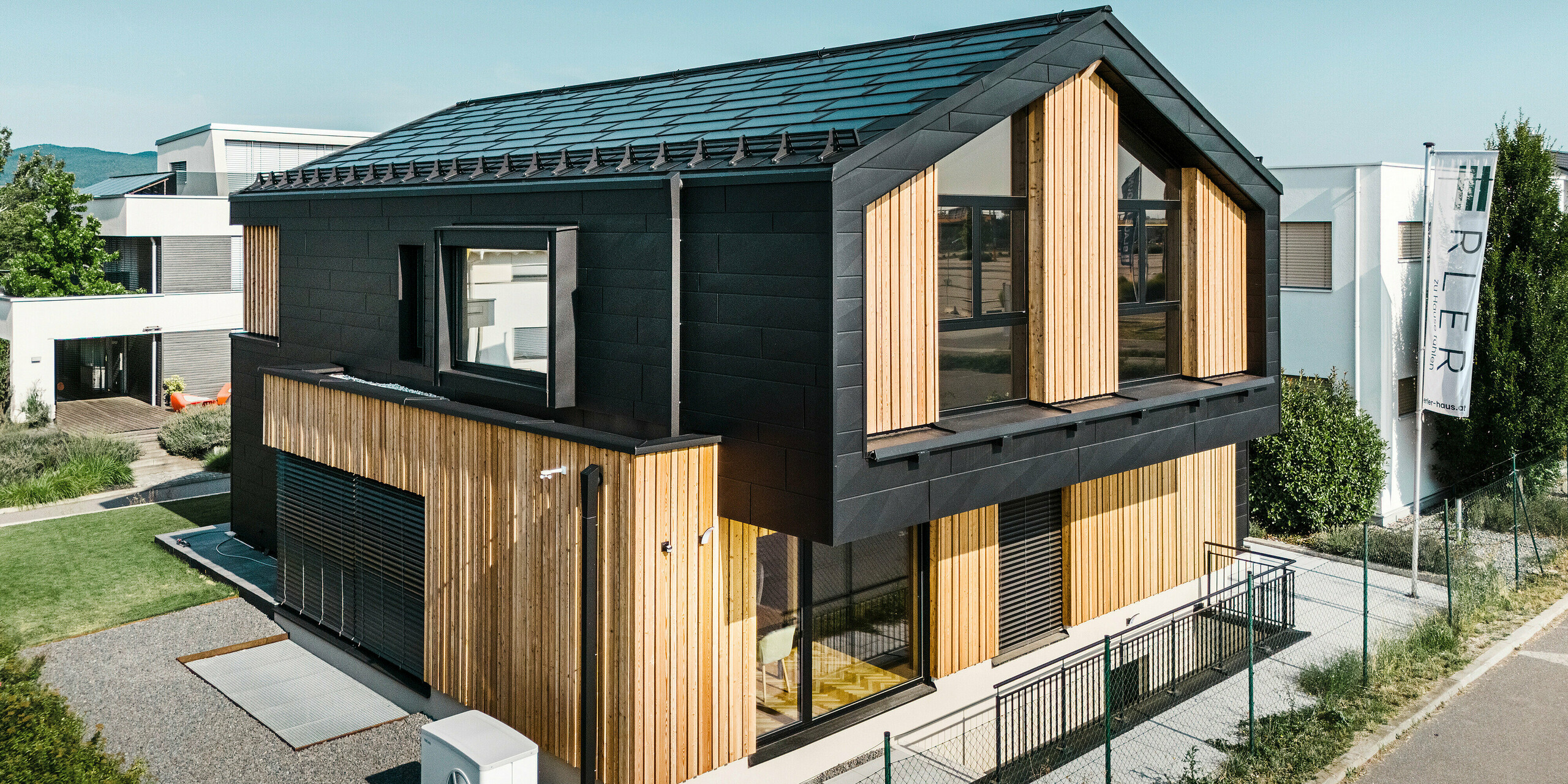 Façade de la maison modèle Autark dans le centre de maisons préfabriquées Blaue Lagune, Vösendorf (AT). Le toit est recouvert de tuiles solaires PREFA noirs et de R.16 avec un revêtement P.10 de haute qualité. La façade est constituée d'une combinaison d'éléments verticaux en bois et de PREFA Siding.X en P.10 noir, qui confère au bâtiment un caractère moderne et durable. Le drapeau de la société Erler Bau GmbH flotte sur le bord droit de l'image.