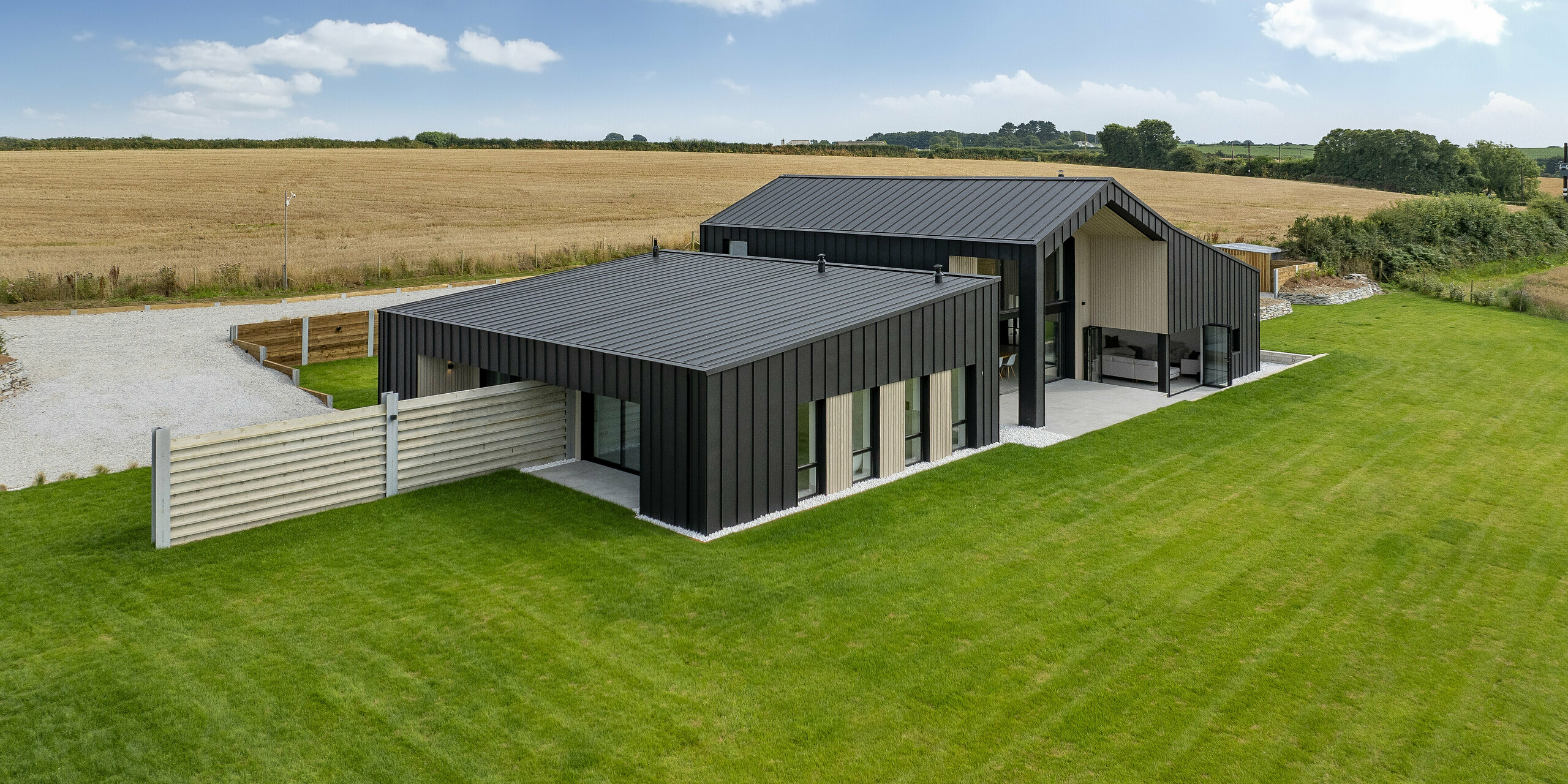 Weitwinkelansicht der Rückseite von ‚The Hide‘ in Cornwall. Das moderne Einfamilienhaus mit PREFALZ Dach- und Fassadenbekleidung aus Aluminium fügt sich harmonisch in die ländliche Umgebung mit vielen Feldern ein. Die anthrazitfarbene Stehfalzdeckung unterstreicht das minimalistische Design und bietet gleichzeitig optimalen Witterungsschutz.