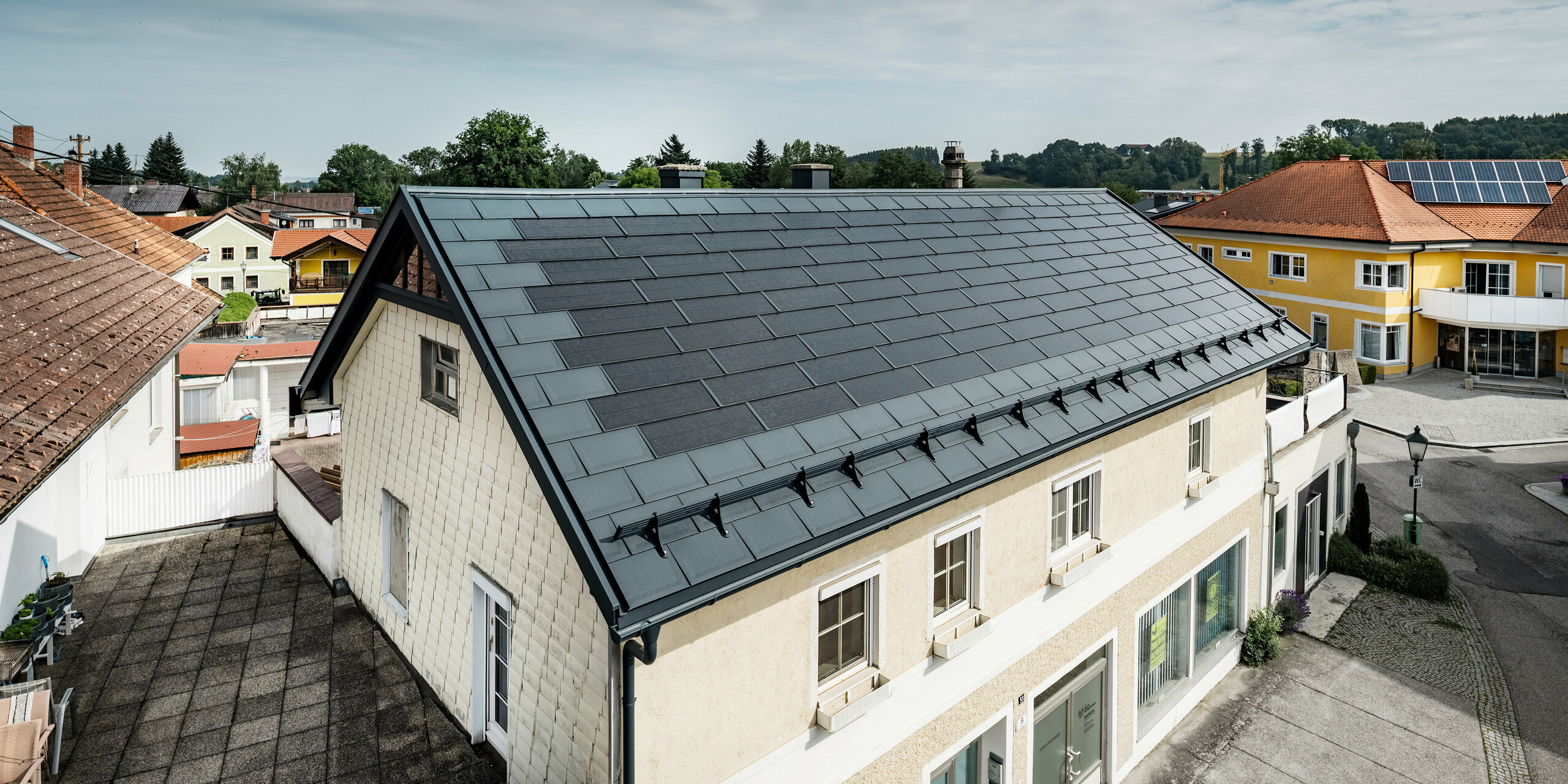 Vue du dessus d’une maison familiale à Mettmach, en Autriche. Le bâtiment le plus ancien a été rénové avec des tuiles solaires PREFA de couleur anthracite. Les systèmes de protection contre la neige et les lignes claires des panneaux solaires soulignent la finition soignée des produits en aluminium PREFA. Le toit de tuiles orange d’un immeuble voisin contraste en arrière-plan. Les modules photovoltaïques sont installés sur le toit de la maison individuelle voisine dans une solution de toit classique, qui montre la diversité des conceptions de toit économes en énergie.