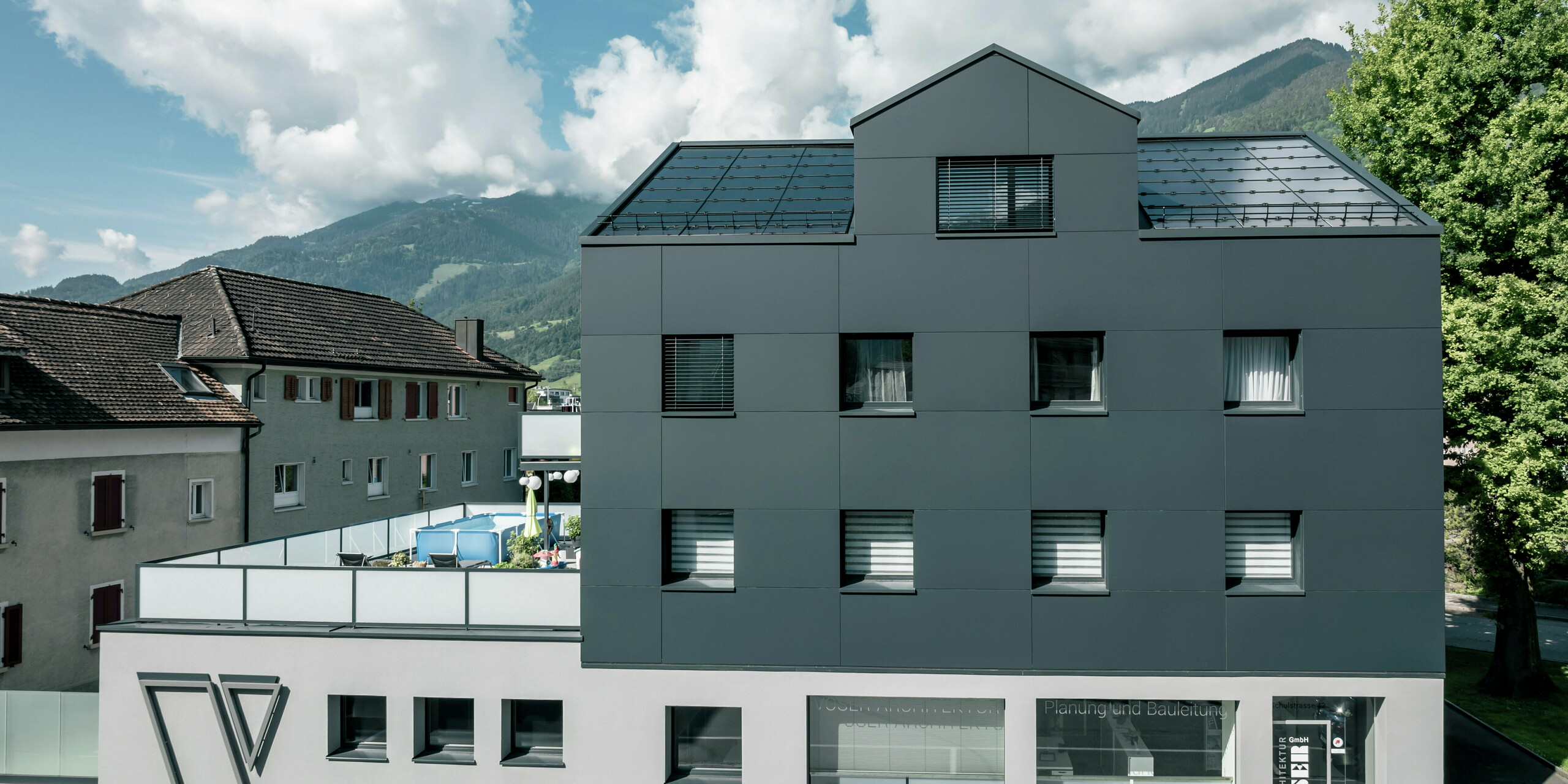 Vue de face d'un bâtiment commercial rénové après rénovation de façade avec des panneaux composites en aluminium PREFABOND en anthracite. La façade claire et de qualité confère au bâtiment un aspect élégant et contemporain. Au rez-de-chaussée se trouvent de grandes fenêtres qui représentent le bureau d'architecture Voser.