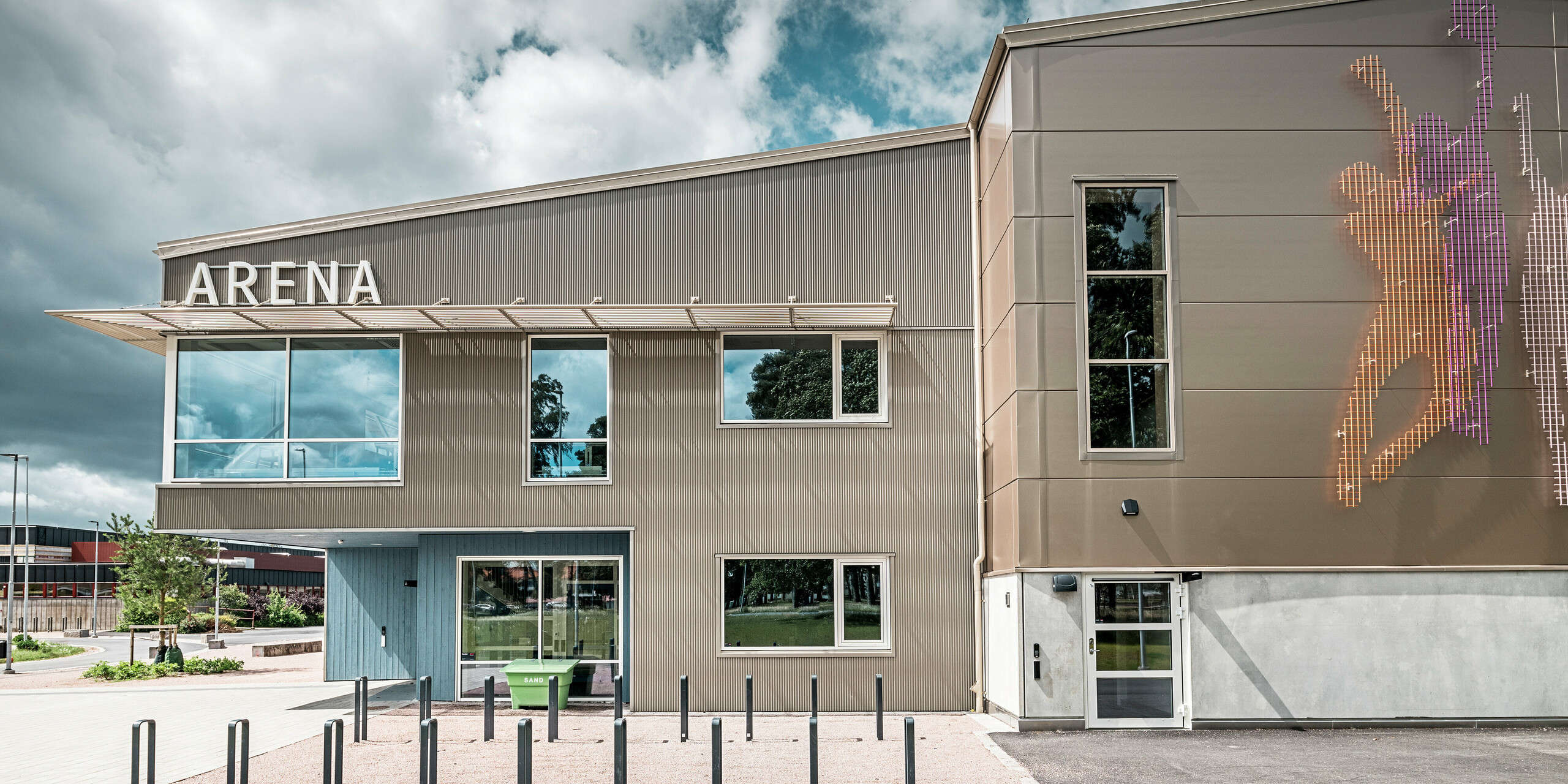 Vue latérale de la zone d'entrée du Park Arena de Värnamo, en Suède, avec une façade PREFA moderne constituée d'un profil triangle en bronze. La façade en aluminium est durable et crée des effets d’ombre et de lumière saisissants. À droite de l'image se trouve une fresque murale de conception artistique qui représente des personnages sportifs et souligne l'atmosphère dynamique de la salle de sport. Les grandes surfaces vitrées laissent entrer beaucoup de lumière dans le bâtiment et soulignent le design contemporain.