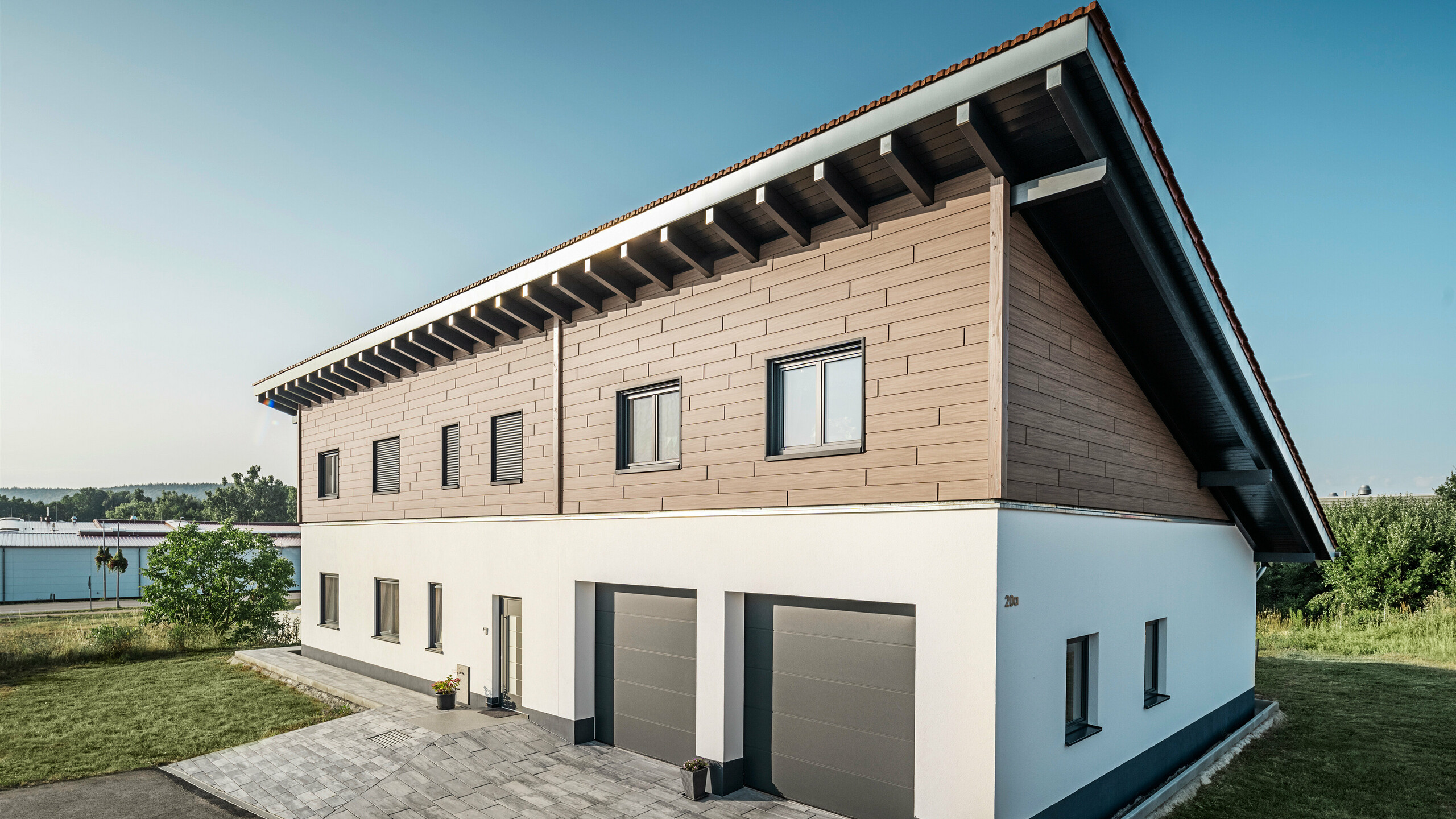 Zicht vanaf de zijkant op een vrijstaand huis met een lessenaarsdak in Bruck. De bovenste verdieping is bekleed met PREFA Sidings in walnoot Brown. Rechts op de foto zijn twee antracietkleurige garagedeuren te zien. De toegangsdeur bevindt zich in het midden van het gebouw. De plint is effen wit. 