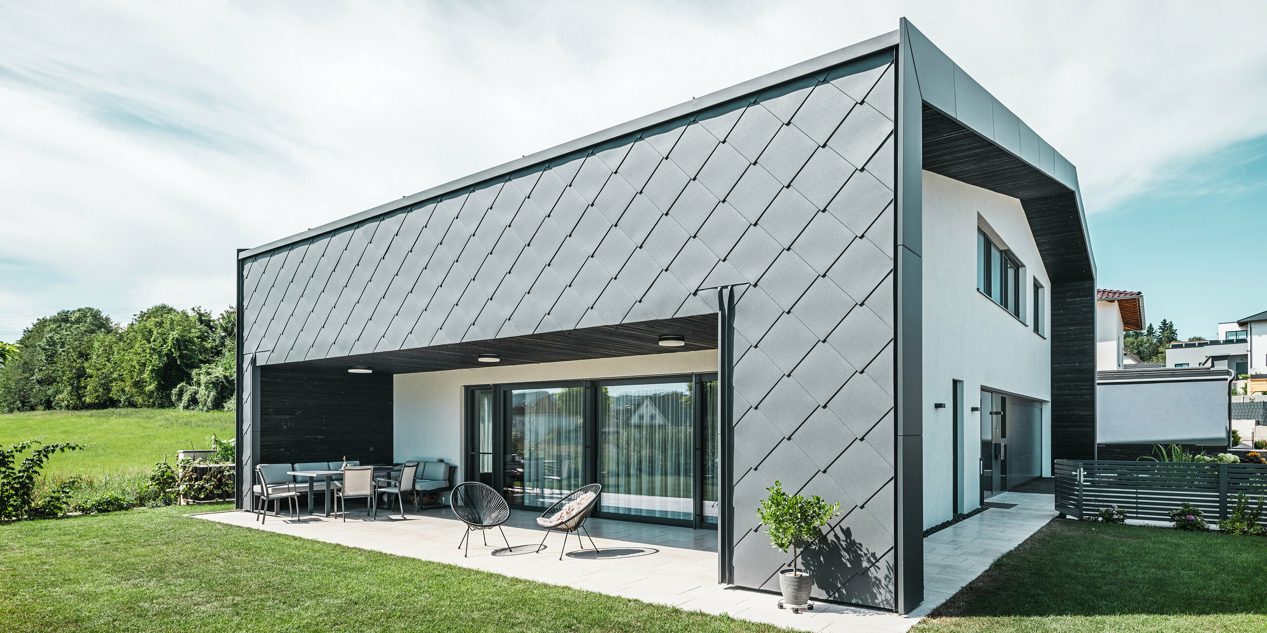 Jardin bien entretenu d'une maison unifamiliale à Aschach. Le bâtiment est élégamment recouvert de losanges de façade 44x44 gris sombre. La terrasse couverte offre un coin salon confortable avec vue sur la pelouse verte luxuriante. Le revêtement élégant en aluminium associé à des surfaces en plâtre blanc garantit une architecture moderne et résistante aux intempéries. De grandes fenêtres laissent entrer beaucoup de lumière du jour à l'intérieur et créent une atmosphère ouverte et lumineuse.