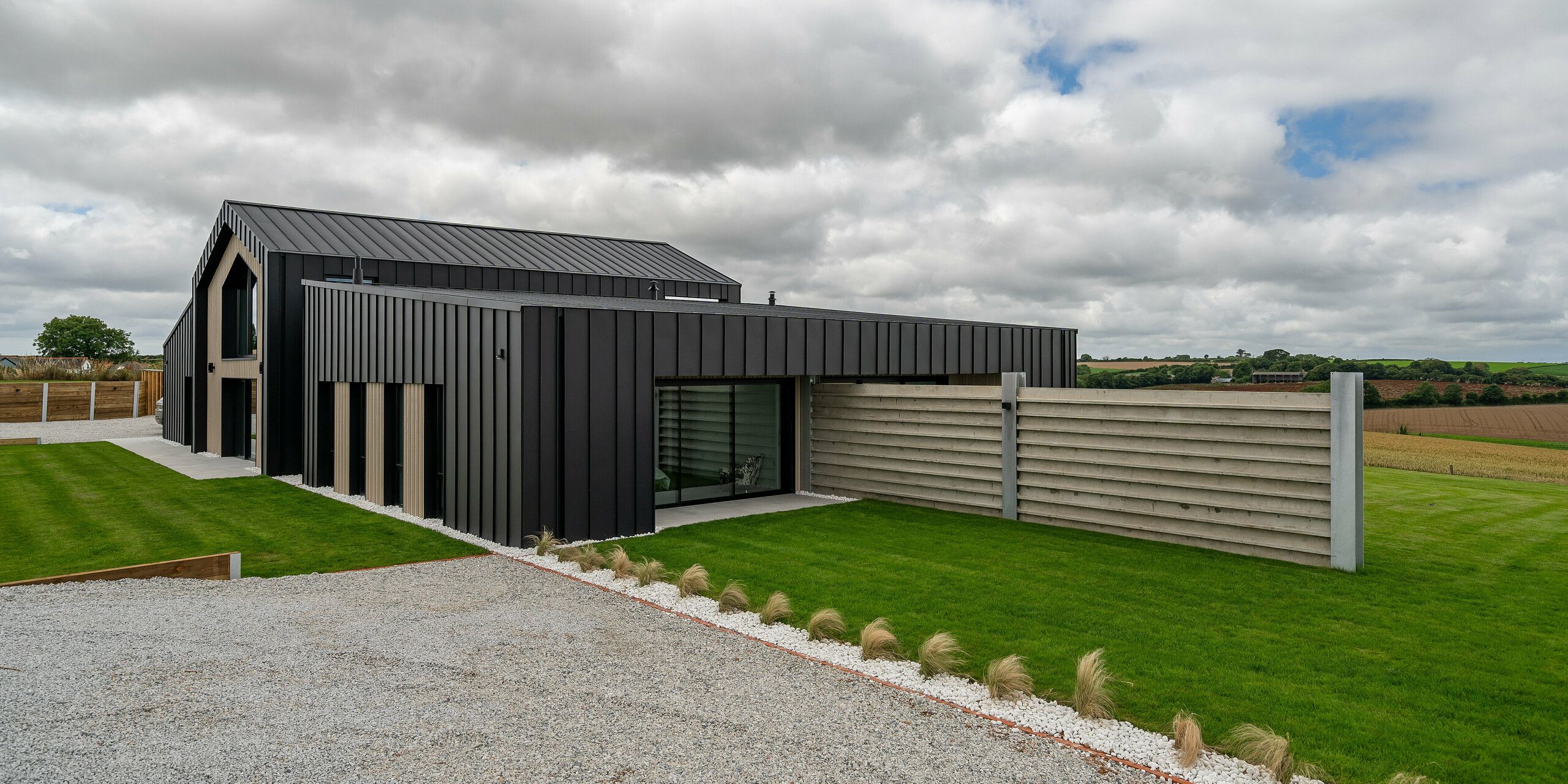 Seitenansicht von ‚The Hide‘ in Cornwall mit PREFALZ Aluminium-Fassade und -Dach in P.10 Anthrazit. Die moderne Stehfalzoptik harmoniert mit der klaren Linienführung des Gebäudes und den umgebenden Naturflächen. Der vorgelagerte Garten und die geschützte Terrasse unterstreichen den funktionalen und stilvollen Charakter des Hauses.