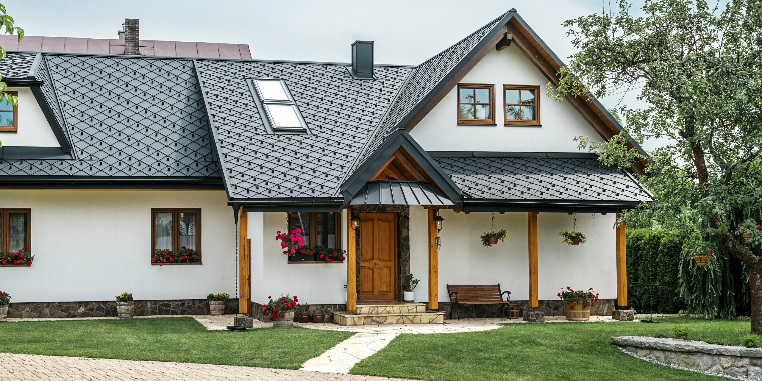 Zone d'entrée d'une maison familiale traditionnelle à Stara Bystrica, Slovaquie, avec un toit en aluminium composé de losanges PREFA 29 × 29 en P.10 noir. Le toit en aluminium de haute qualité au look élégant comprend des fenêtres de toit inclinées et une véranda avec auvent recouvert de PREFALZ. Les produits en aluminium à la fois élégants et robustes confèrent à la maison une apparence contemporaine et accueillante. Le jardin bien entretenu et les compositions florales soulignent le bon entretien du bâtiment.