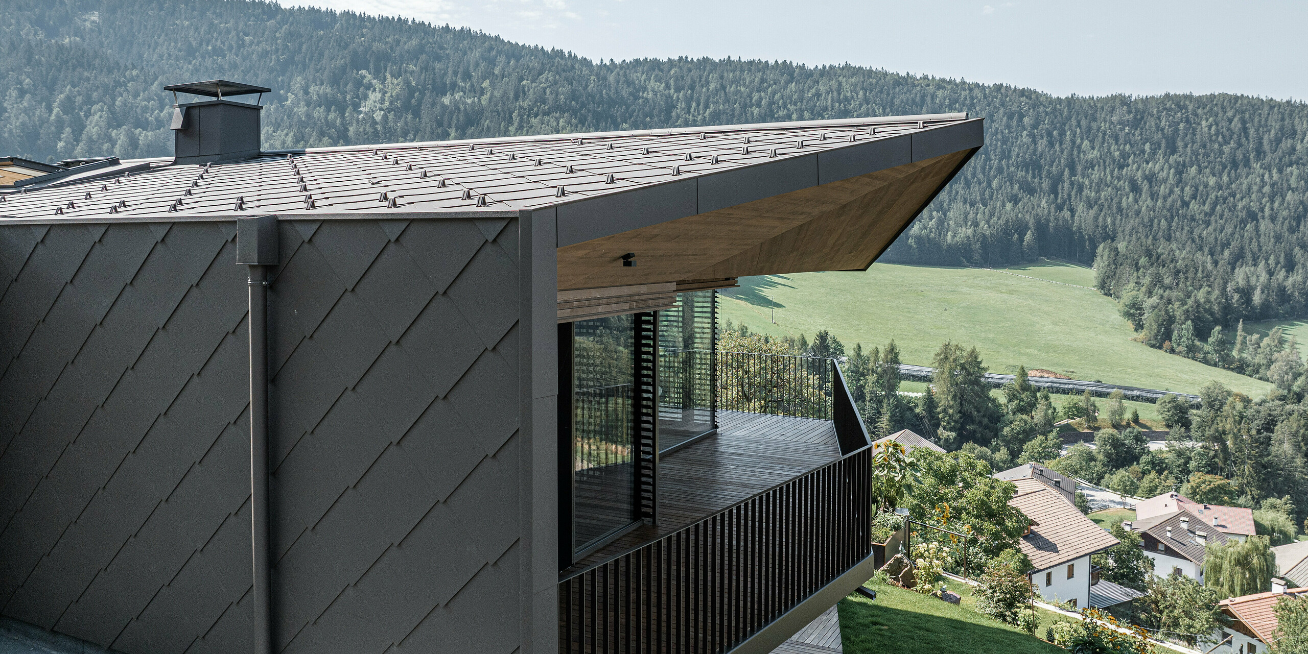 Nahaufnahme einer Ecke einer modernisierten Doppelhaushälfte in Mölten, Südtirol, mit dunkelbraunen Aluminiumrauten von PREFA auf dem Dach und an der Fassade. Das Design integriert eine große überdachte Terrasse mit Holzgeländer, die einen atemberaubenden Blick auf die umliegende grüne Landschaft und die fernen Hügel bietet. Der sorgfältig gestaltete Übergang zwischen den Materialien unterstreicht die harmonische Verbindung von Natur und moderner Architektur. Das ursprünglich Anfang der 1990er Jahre errichtete Gebäude wurde im Rahmen der Renovierung großzügig umgebaut und renoviert.