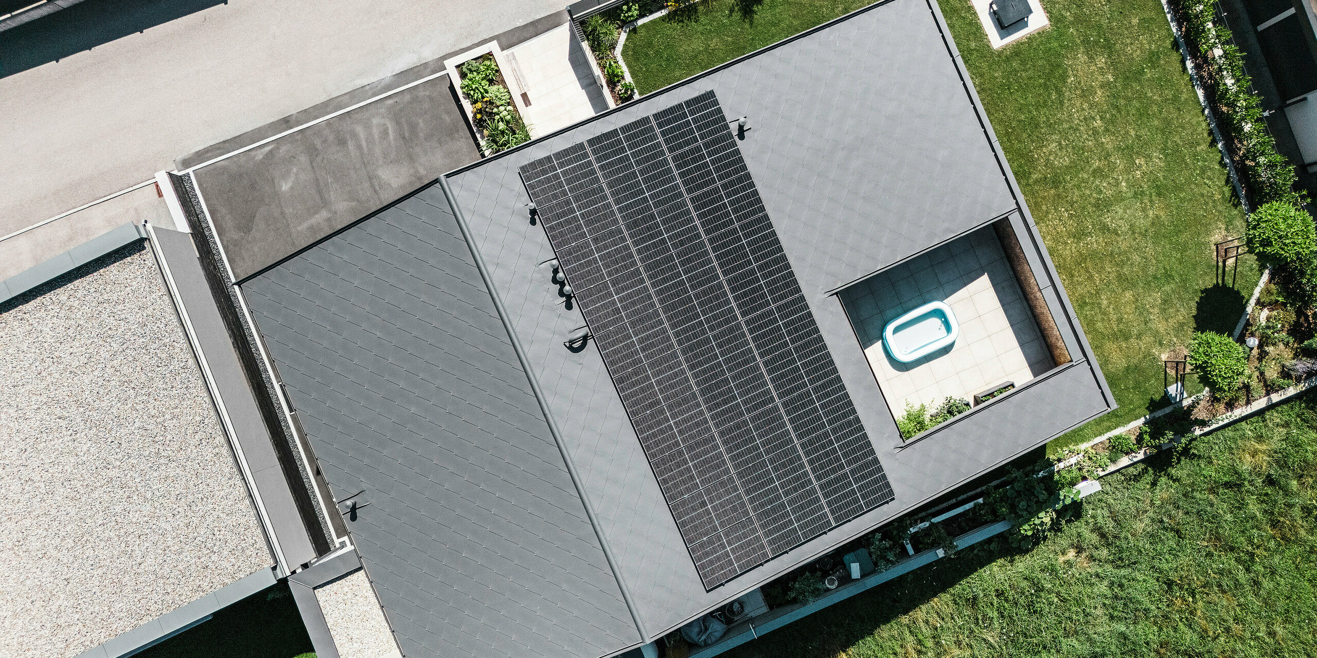 Vue aérienne d'une maison familiale moderne à Aschach avec des losanges 44x44 PREFA gris sombre en qualité P.10. La surface du toit est couverte d'un système photovoltaïque à grande échelle et intègre un atrium ouvert avec une petite piscine. La combinaison de diamants en aluminium durables et d'un design sophistiqué garantit une protection optimale contre les intempéries et une apparence attrayante. Le jardin environnant donne à la maison un aspect soigné.