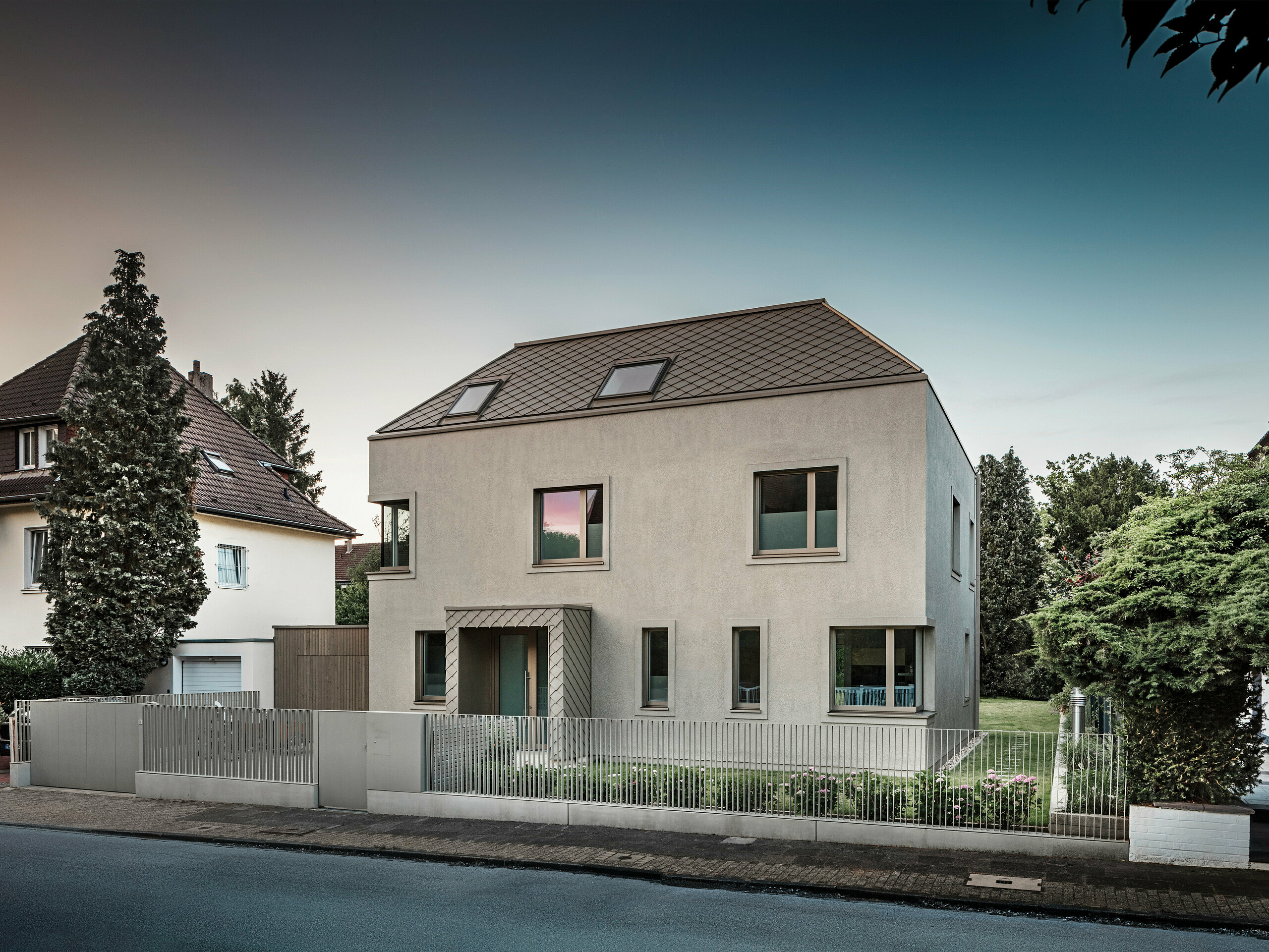 Une maison individuelle moderne avec un toit en croupe et un revêtement de toiture et de façade en aluminium couleur bronze de PREFA. La maison est située dans un quartier résidentiel calme, entourée de jardins bien entretenus et d'autres maisons individuelles. Au premier plan, vous pouvez voir une clôture basse qui sépare la propriété de la rue. L'architecture de la maison se caractérise par des lignes épurées et de grandes fenêtres qui laissent entrer beaucoup de lumière naturelle dans les intérieurs.