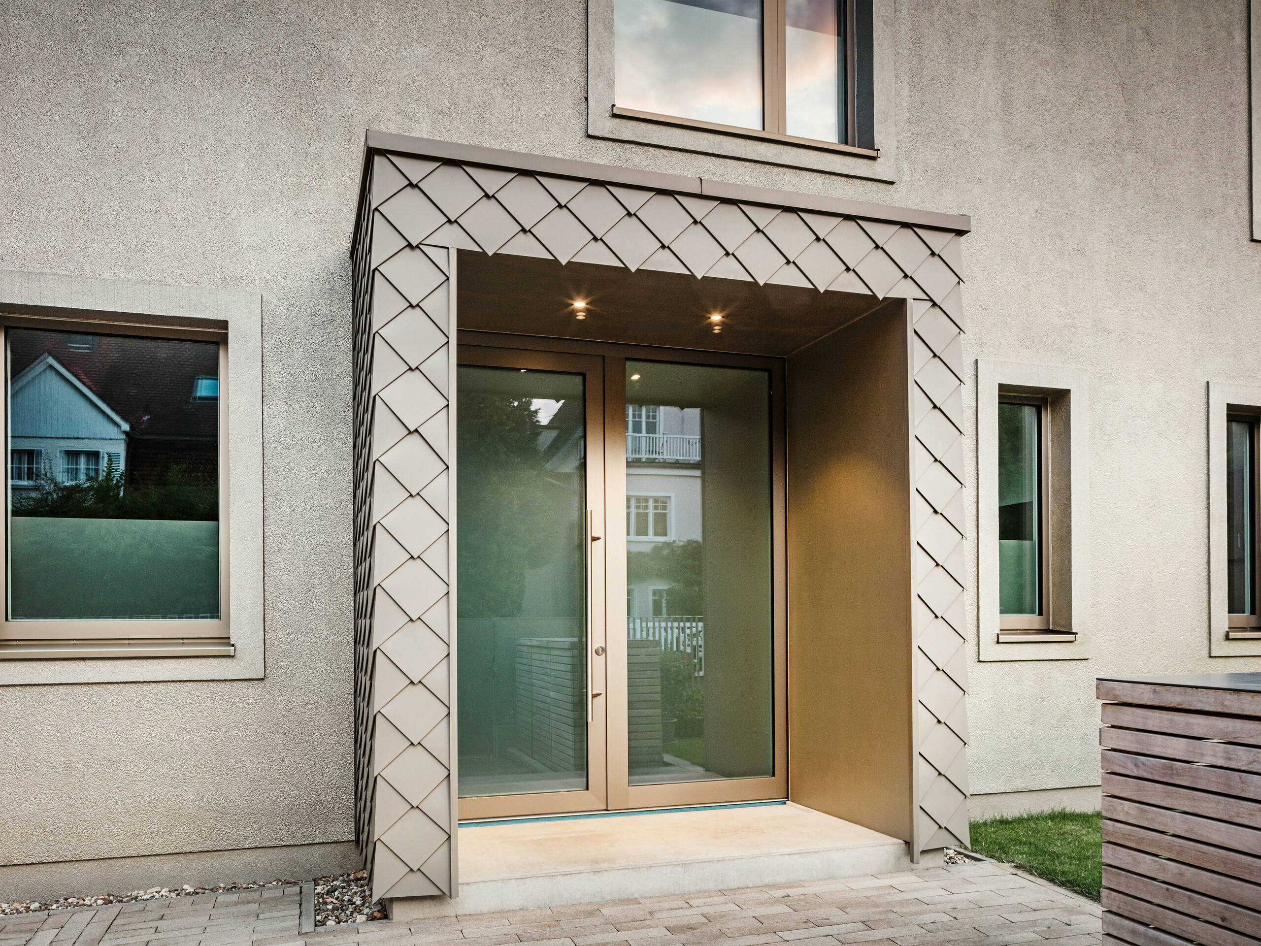 Vue détaillée de l'entrée d'une maison individuelle moderne avec revêtement en aluminium couleur bronze de PREFA. L'entrée est recouverte et revêtue d'élégants losanges en aluminium, donnant à la zone un aspect moderne et élégant. Il y a de grandes fenêtres à gauche et à droite de la porte d'entrée, créant une atmosphère lumineuse et accueillante.