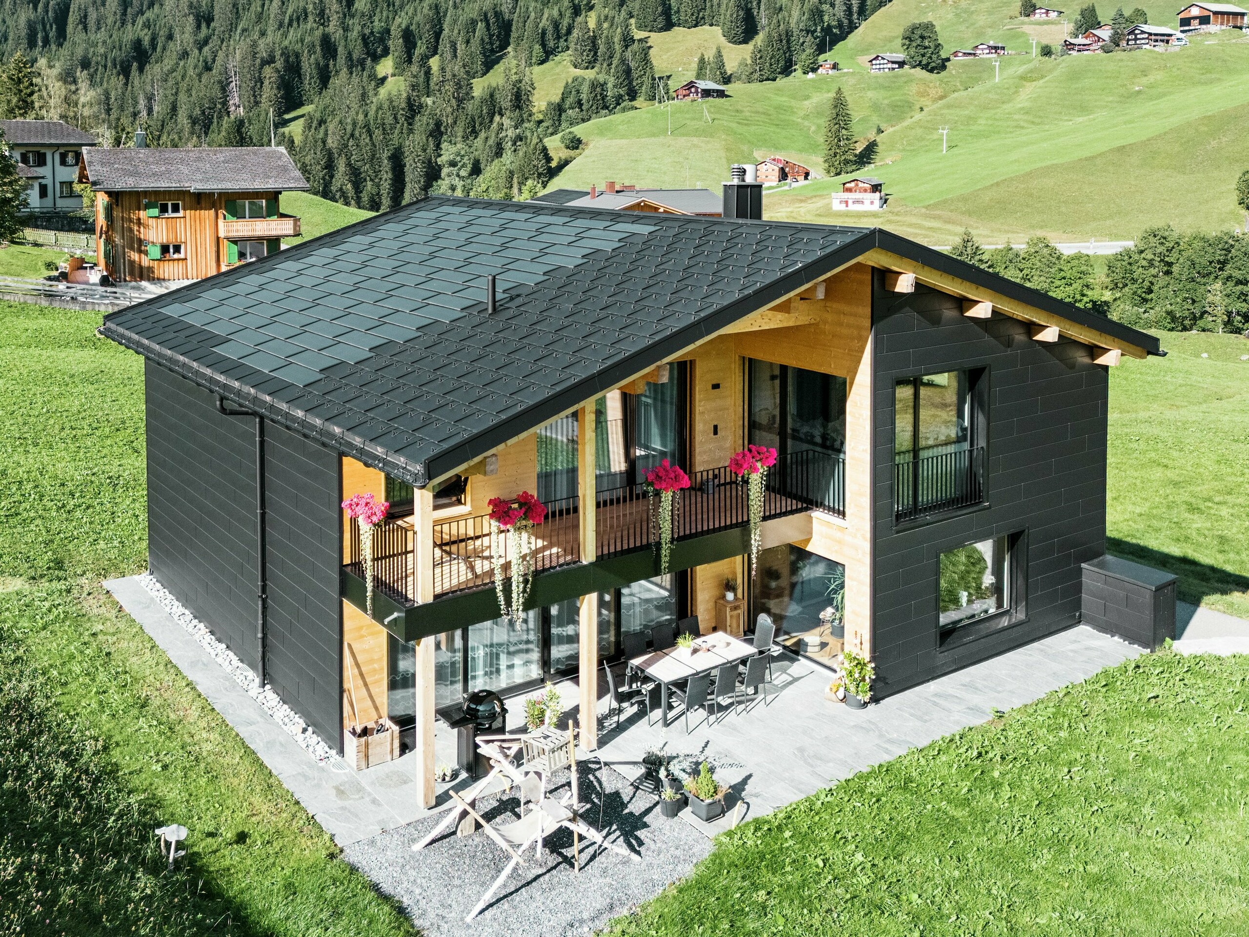 Cette élégante maison individuelle située dans les environs vallonnés d'Ascharina (Suisse) montre comment l'aluminium et le bois peuvent être parfaitement combinés. Les robustes panneaux de façade PREFA Siding.X en noir P.10 s'harmonisent avec le toit composé de panneaux solaires PREFA et de panneaux de toiture R.16 dans le même revêtement de couleur. L'enveloppe en aluminium durable protège le bâtiment de manière fiable contre les défis du climat alpin.