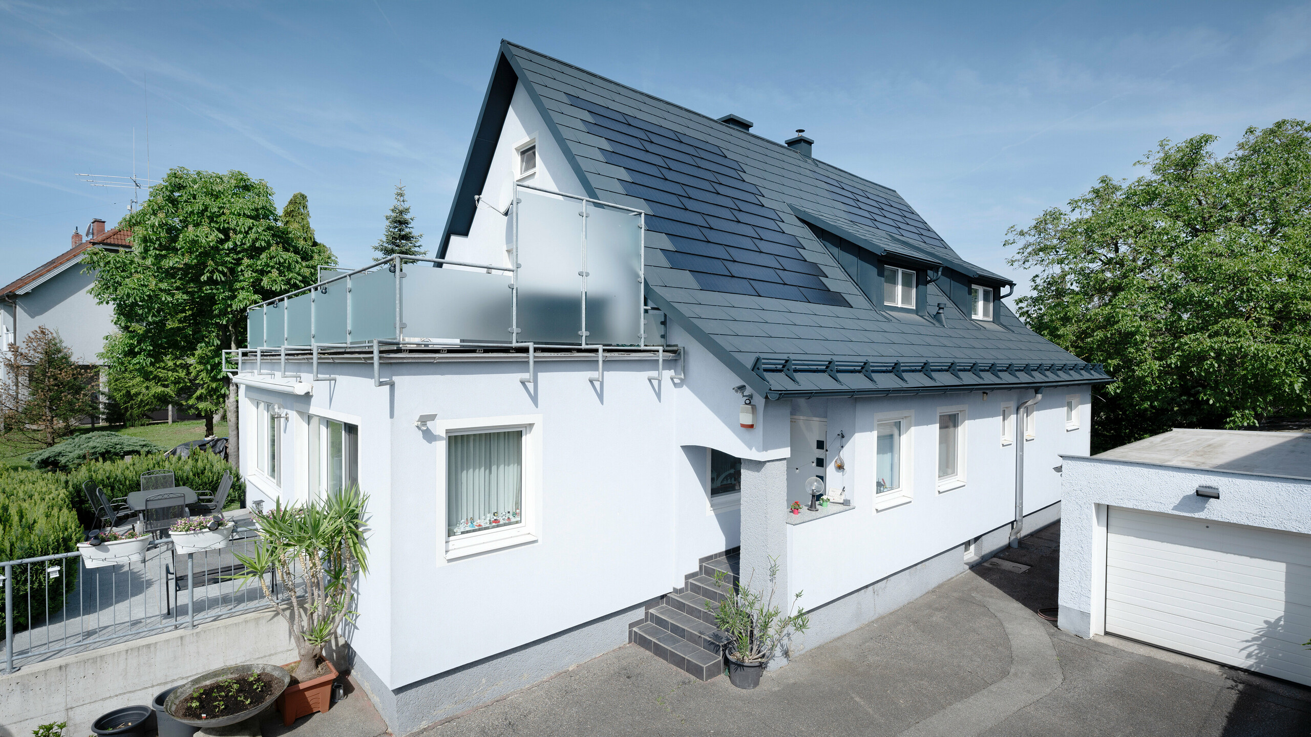 Dakrenovatie van een vrijstaand huis in Leonding  met PREFA-zonnedakpanelen in antraciet. Op de foto is het gerenoveerde huis van opzij te zien met een duidelijk zicht op het dak en de geïntegreerde zonnepanelen. Deze bieden een duurzame en energie-efficiënte manier om stroom op te wekken. In deze woning wekken de zonnepanelen een stroomopbrengst van 10,6 kWp op. De moderne, duurzame en weerbestendige PREFA-materialen dragen bij aan een esthetische en functionele dakconstructie die voldoet aan de nieuwste normen voor duurzaam bouwen.