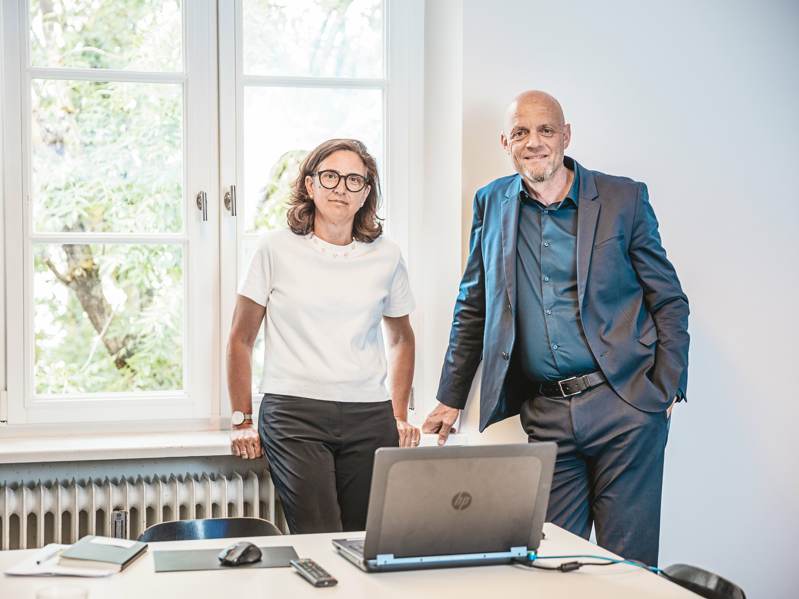 Patricia Ezcura et Gerold Heugenhauser de WWA Architekten de Munich, les esprits créatifs derrière le projet résidentiel d'Utting am Ammersee. Les deux se tiennent dans un bureau derrière une table avec un ordinateur portable et des cahiers et devant une grande fenêtre. Leur planification et conception innovantes du complexe immobilier moderne sur le site Schmucker avec des produits PREFA durables tels que le R.16 se caractérisent par une intégration harmonieuse dans l'environnement et une architecture esthétique.