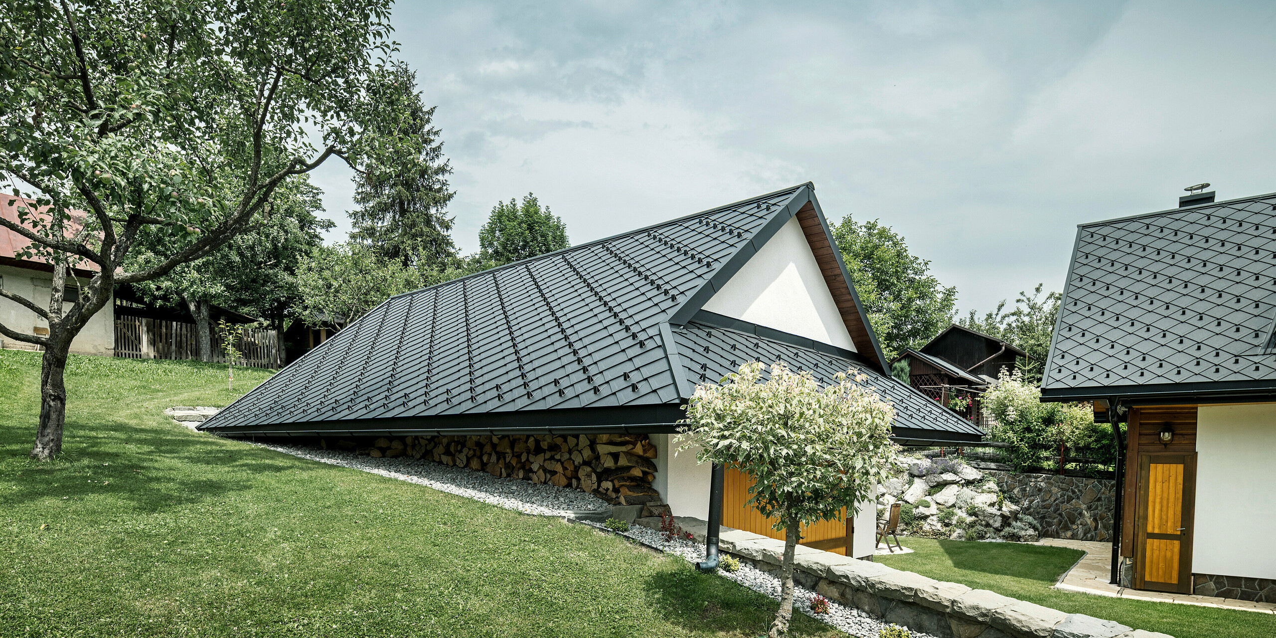 Dépendance d'une maison familiale traditionnelle à Stara Bystrica, Slovaquie, avec un toit en losanges PREFA 29 × 29 en P.10 noir. Le toit en aluminium robuste protège le bâtiment et s'intègre harmonieusement dans l'environnement verdoyant et rural. Au premier plan, vous pouvez voir une pelouse bien entretenue et un arbre, donnant à l'ensemble une atmosphère idyllique et accueillante.