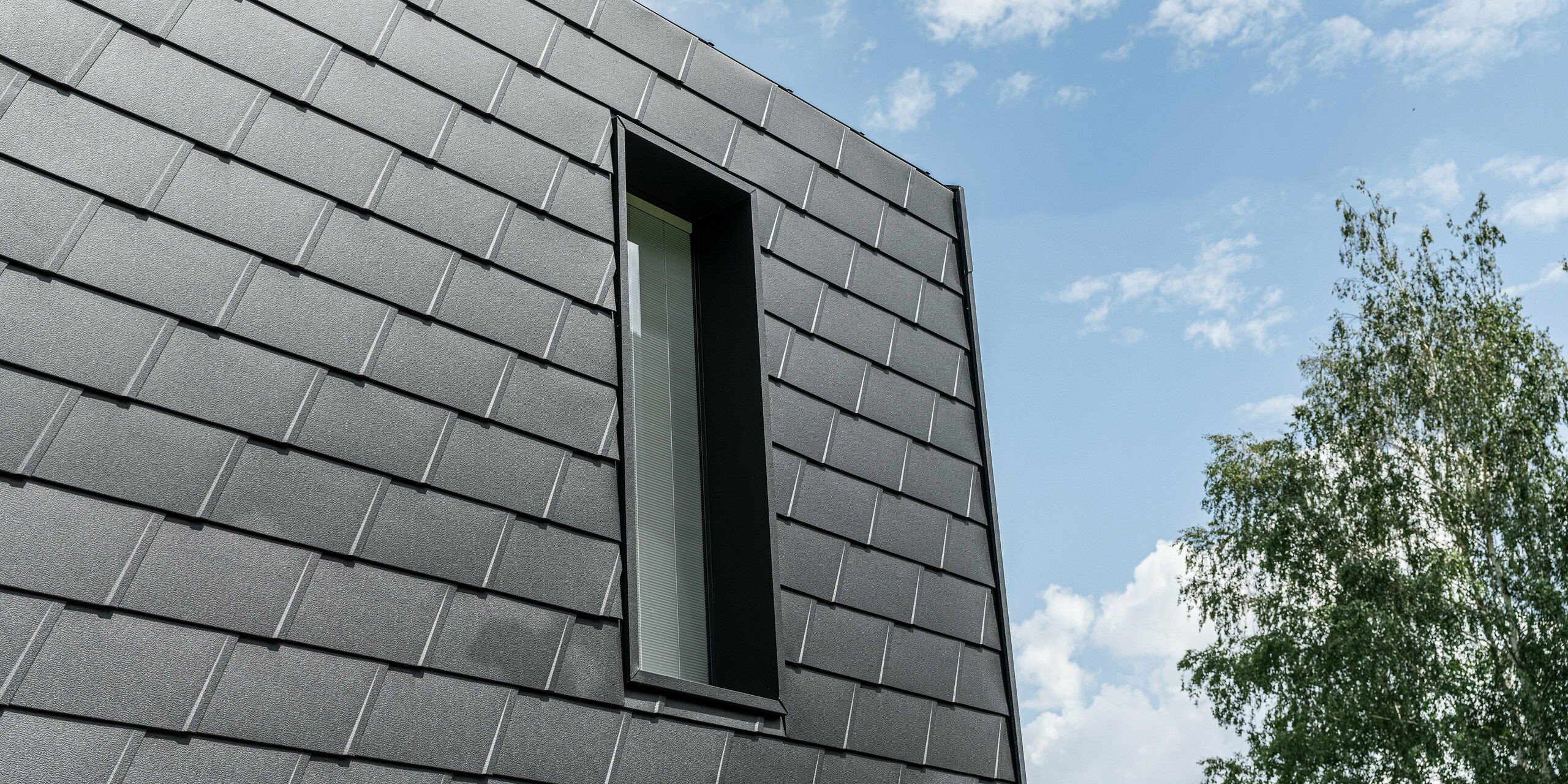 Detailansicht einer Fassade mit PREFA Wandschindeln in Schwarz, die ein schlankes, vertikales Fenster einrahmt. Die Schindeln bieten eine strukturierte, robuste Oberfläche, die ästhetisch mit der natürlichen Umgebung und dem blauen Himmel kontrastiert.