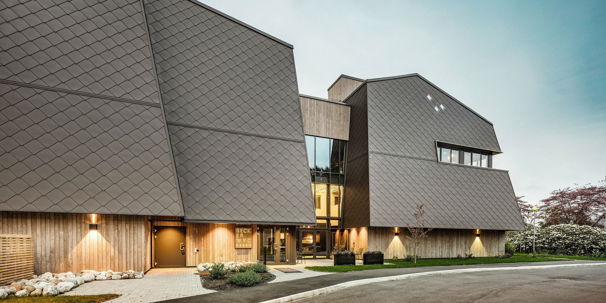 Entrée de l'hôtel-boutique Beckerwyc House à Bekkjarvik, Norvège. Le bâtiment au look futuriste est habillé de losanges de façade PREFA 44 × 44 en noyer foncé, qui forment une coque protectrice qui s'appuie sur l'environnement rocheux. Les éléments de façade inclinés et brun foncé constituent une déclaration architecturale saisissante. L'entrée est entourée d'un éclairage naturel et d'éléments paysagers qui soulignent le design moderne de l'hôtel.