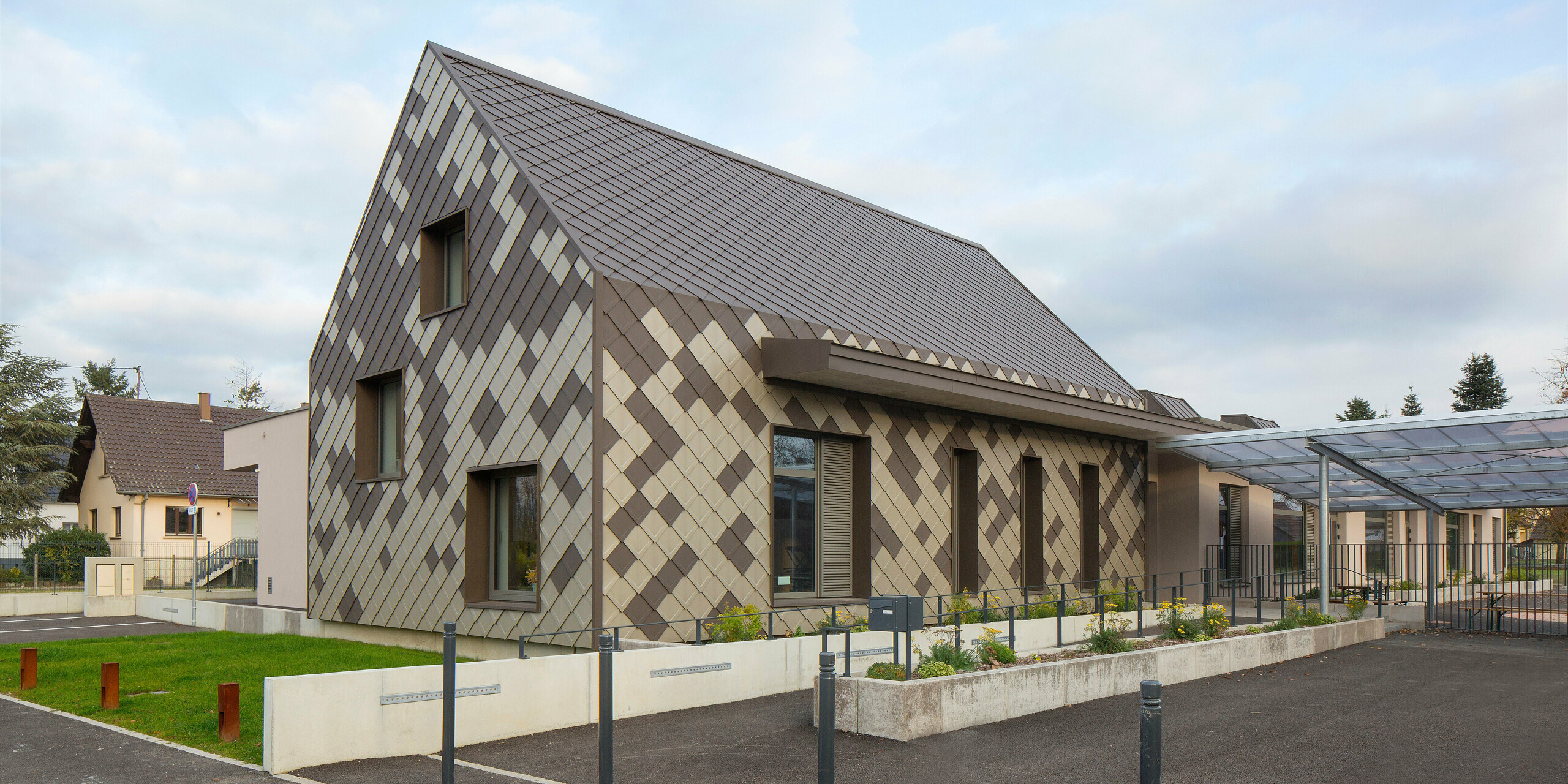 [Translate to Français:] Vue depuis la rue du périscolaire de Sessenheim. Le bâtiment présente une remarquable coque en aluminium dans les couleurs brun et bronze.