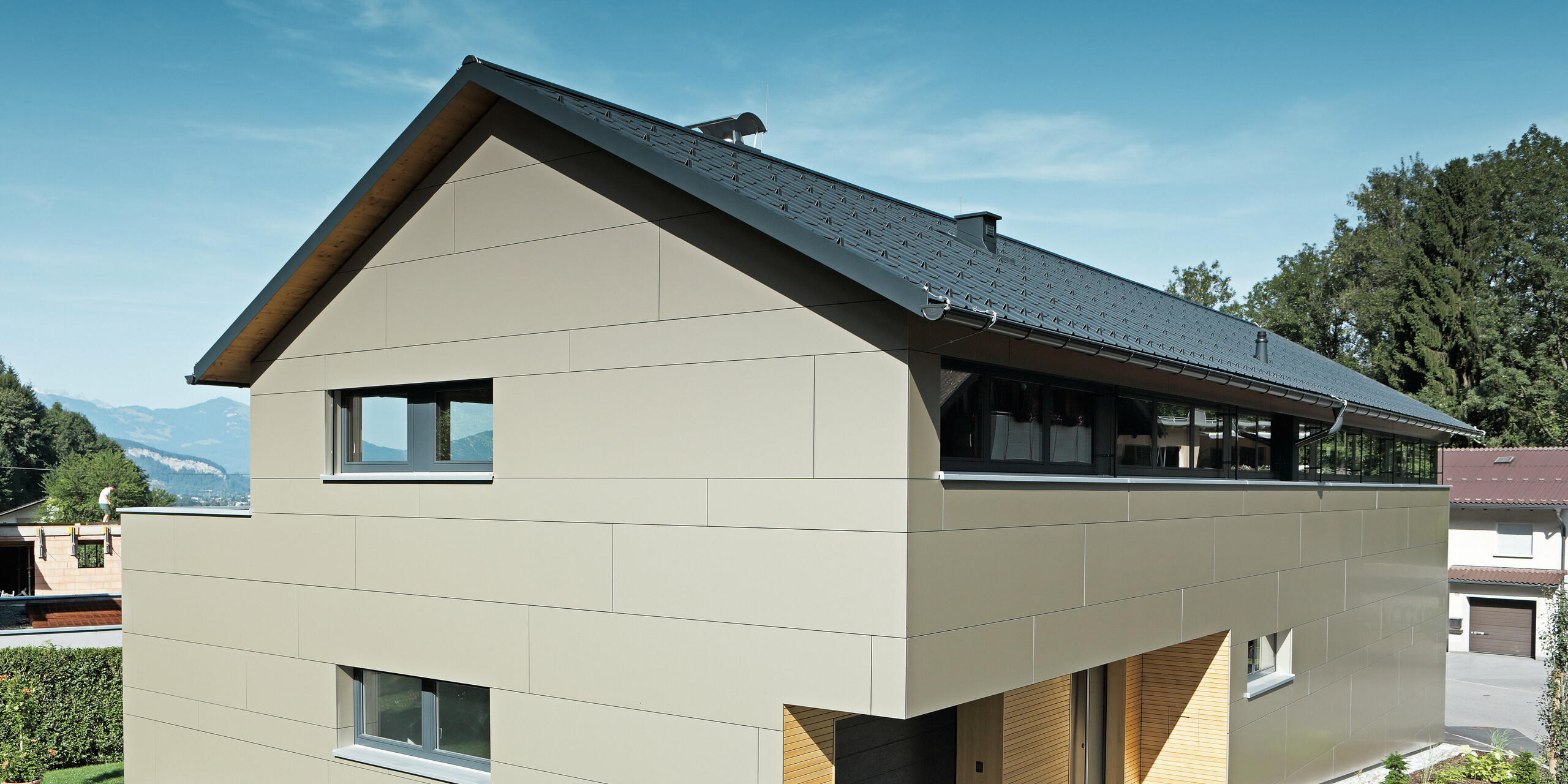 Elegantes Einfamilienhaus in Hohenems mit PREFA Komplettsystem: Die Fassade aus Aluminium Verbundplatten in Bronzemetallic und das Dach aus PREFA Dachplatten in P.10 Anthrazit fügen sich nahtlos in die umgebende Landschaft ein. Die klare, strukturierte Architektur mit dem sichtbaren Holzunterdach, kombiniert mit der robusten, wartungsarmen Aluminiumverkleidung, bietet einen nachhaltigen und ästhetischen Wohnraum, eingebettet in die natürliche Umgebung mit Bergblick im Hintergrund.