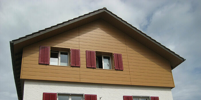 Habillage du pignon d’une maison classique avec toit à deux pans. Le pignon est habillé à l’aide de Sidings PREFA couleur chêne naturel posés à l’horizontale. Les fenêtres disposent de volets de couleur rouge.