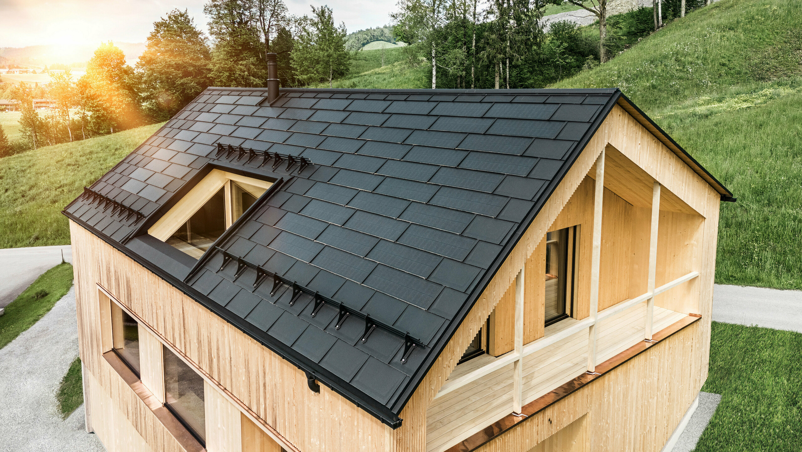 Blick auf ein modernes, nachhaltiges Holzhaus in Egg mit einem PREFA Solardach, das durch seine dunkle, strukturierte sowie elegante Erscheinung auffällt. Die Solarmodule sind strategisch zwischen den klassischen Dachplatten R.16 platziert, um eine optimale Sonneneinstrahlung zu nutzen und sich dabei ästhetisch in das Gesamtdesign einzufügen. Das Haus steht im Einklang mit seiner grünen Umgebung, während die warme Abendsonne die natürliche Holztextur der Fassade unterstreicht und die solide Bauweise betont. Ein harmonisches Beispiel für umweltbewusstes Bauen und Energieeffizienz.