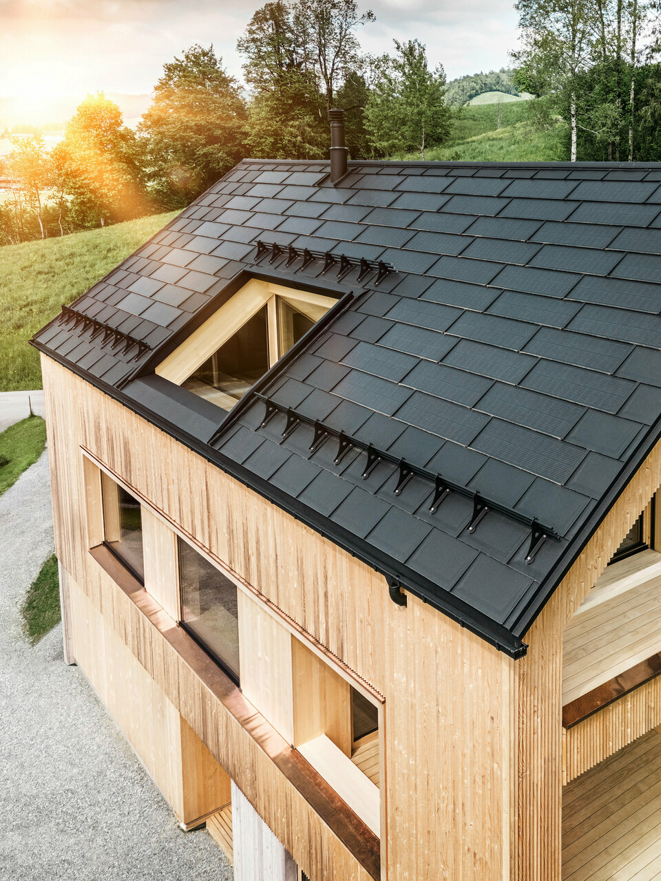 Blick auf ein modernes, nachhaltiges Holzhaus in Egg mit einem PREFA Solardach, das durch seine dunkle, strukturierte sowie elegante Erscheinung auffällt. Die Solarmodule sind strategisch zwischen den klassischen Dachplatten R.16 platziert, um eine optimale Sonneneinstrahlung zu nutzen und sich dabei ästhetisch in das Gesamtdesign einzufügen. Das Haus steht im Einklang mit seiner grünen Umgebung, während die warme Abendsonne die natürliche Holztextur der Fassade unterstreicht und die solide Bauweise betont. Ein harmonisches Beispiel für umweltbewusstes Bauen und Energieeffizienz.