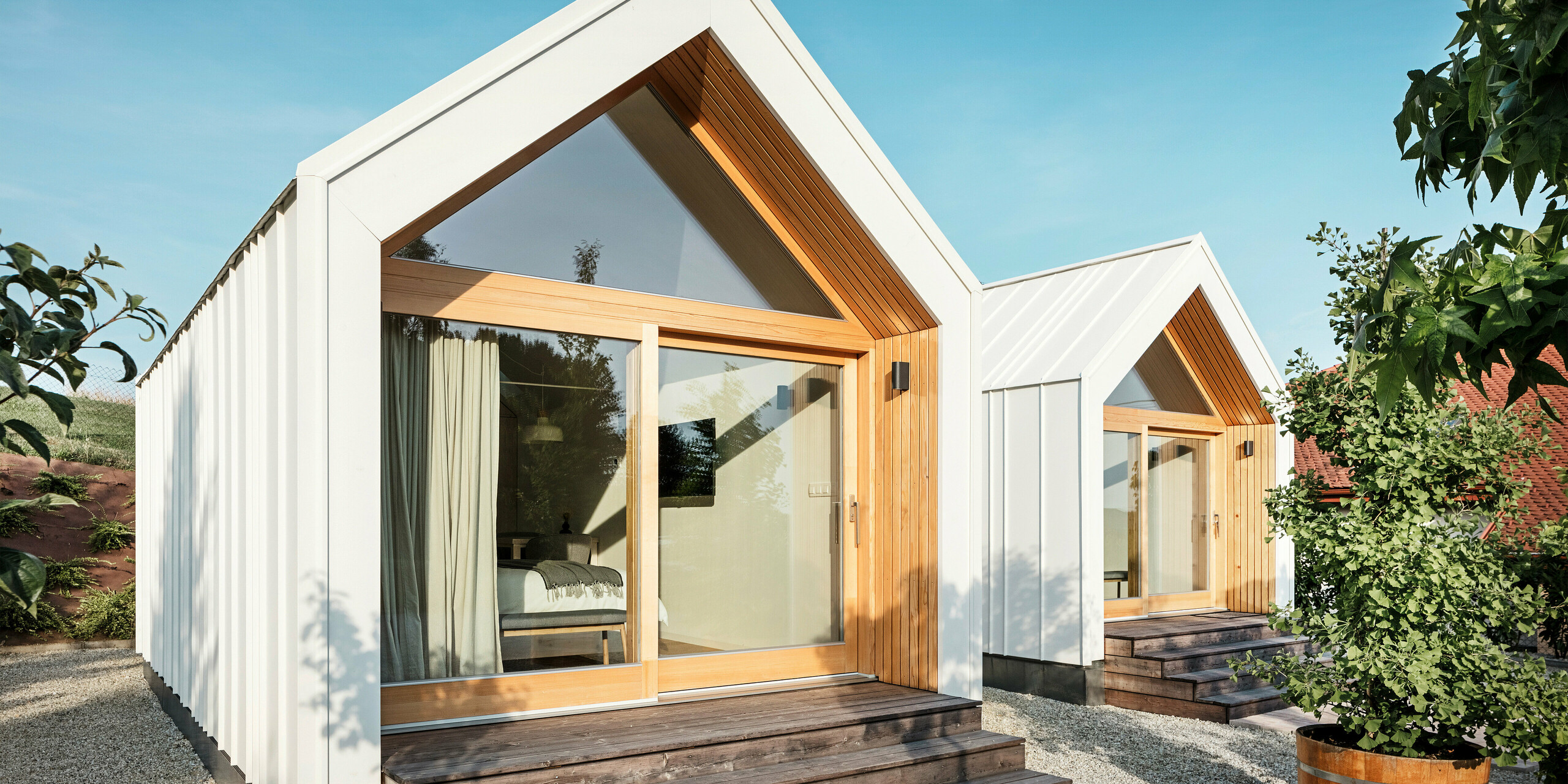 Vue de face des deux petites maisons de la résidence d'hôtes « Pri Momi » à Kančevci, en Slovénie, avec de grandes façades vitrées, conçues avec un toit et un revêtement de façade PREFALZ en blanc pur P.10 et bois naturel. Les micro-maisons minimalistes comportent de larges marches en bois menant aux entrées, offrant une vue directe sur les intérieurs confortables. Les lignes claires et la combinaison de l'aluminium et du bois soulignent le design moderne et accueillant du logement de vacances.