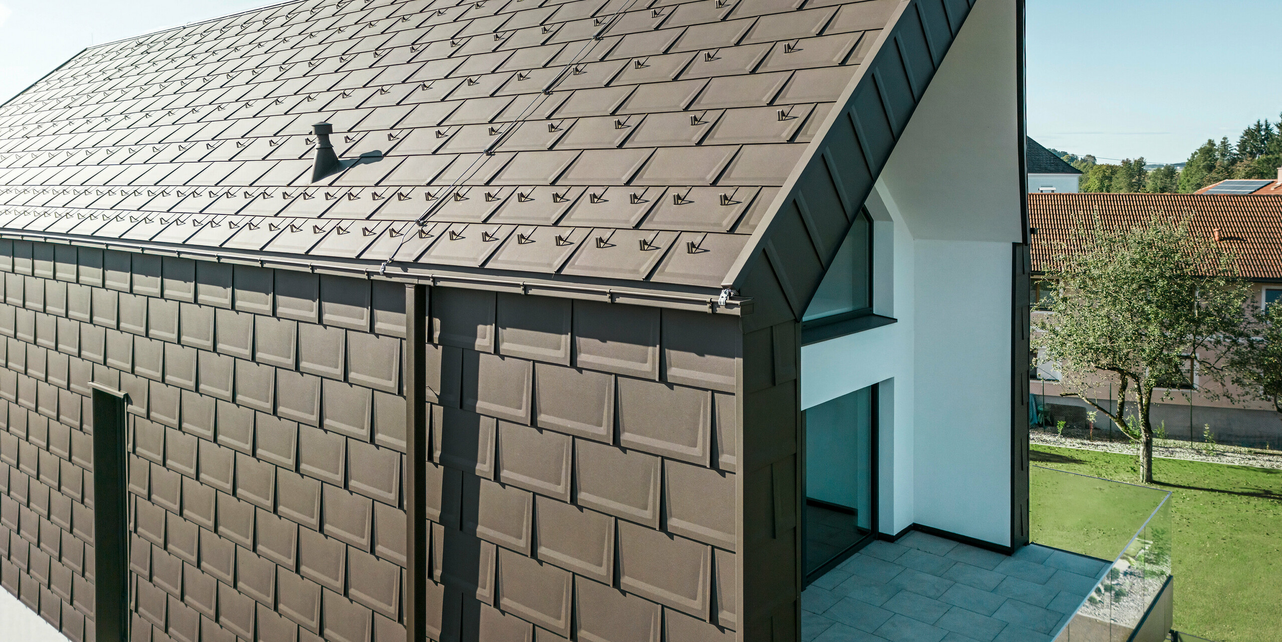 Eine perspektivische Ansicht von Dach und Fassade eines modernen Einfamilienhauses in Neukirchen, Österreich. Die PREFA R.16 Dachplatte im Braunton P.10 bietet eine ästhetische und funktionale Dacheindeckung, die sich perfekt in die architektonische Gestaltung des Hauses einfügt. Der scharfe First des Daches und die präzise Anordnung der Dachplatten zeigen die Qualität von PREFA. Die kleinformatigen Aluminiumprodukte fügen sich perfekt in die weiß verputzte Fassade und die umgebende grüne Landschaft ein.