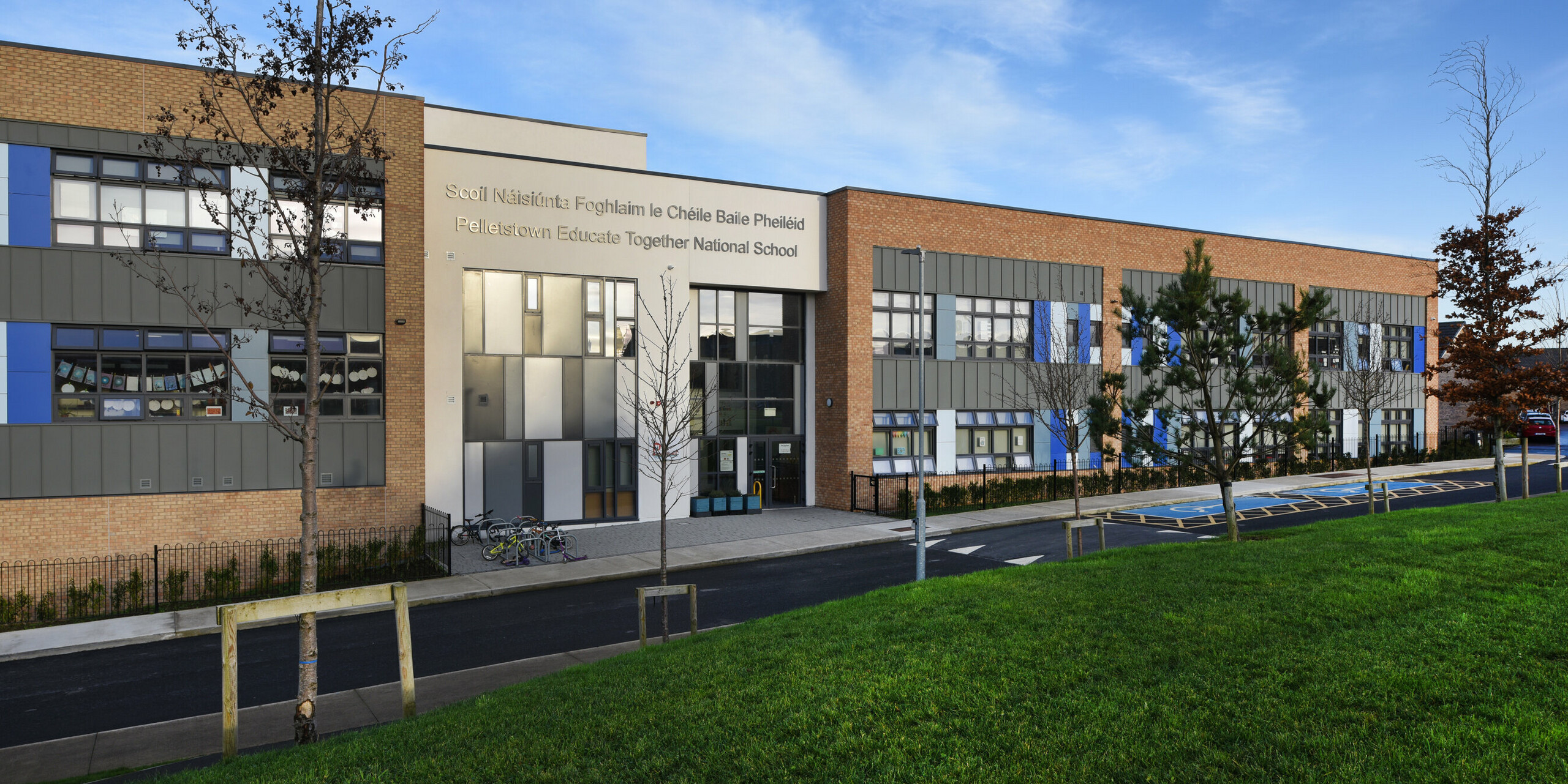 Vue de face de l'école nationale Pelletstown Educate Together à Dublin avec façade moderne PREFALZ en P.10 gris souris. La combinaison harmonieuse de l’aluminium, des briques et du crépi souligne la conception durable et élégante du bâtiment scolaire. Un espace vert bien entretenu avec des arbres au premier plan souligne l'engagement en faveur de la durabilité - tout comme les vélos devant l'entrée.