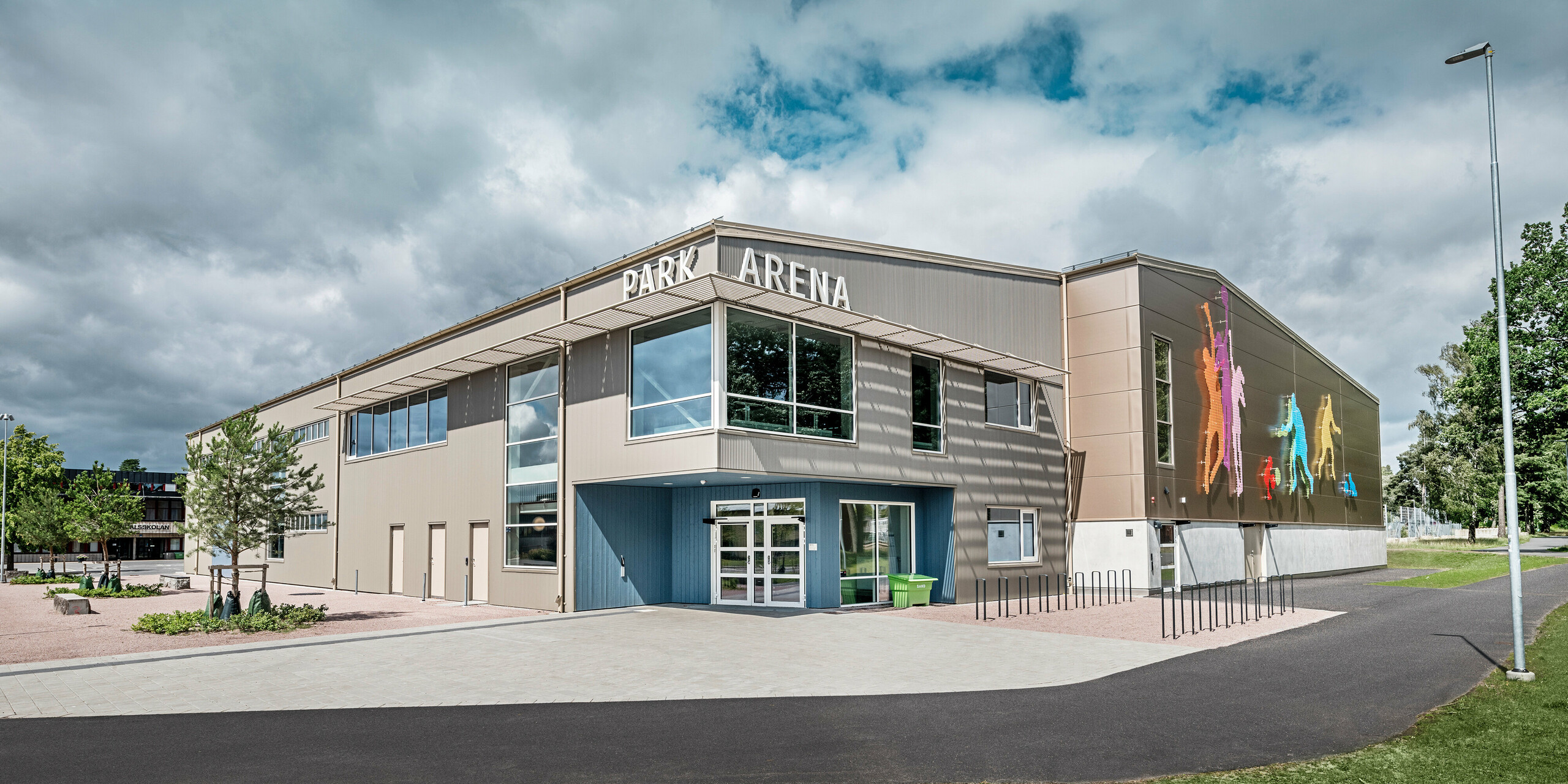 Vue de face du Park Arena de Värnamo, en Suède, avec une façade PREFA moderne composée de profilés triangle en bronze. La façade en aluminium offre une solution durable et résistante aux intempéries qui se distingue par des effets d'ombre et de lumière saisissants. À droite, une fresque murale artistique représente des personnages sportifs colorés qui soulignent le caractère dynamique de la salle de sport. Les grandes fenêtres laissent entrer beaucoup de lumière naturelle dans le bâtiment et renforcent le design moderne de l'arène.