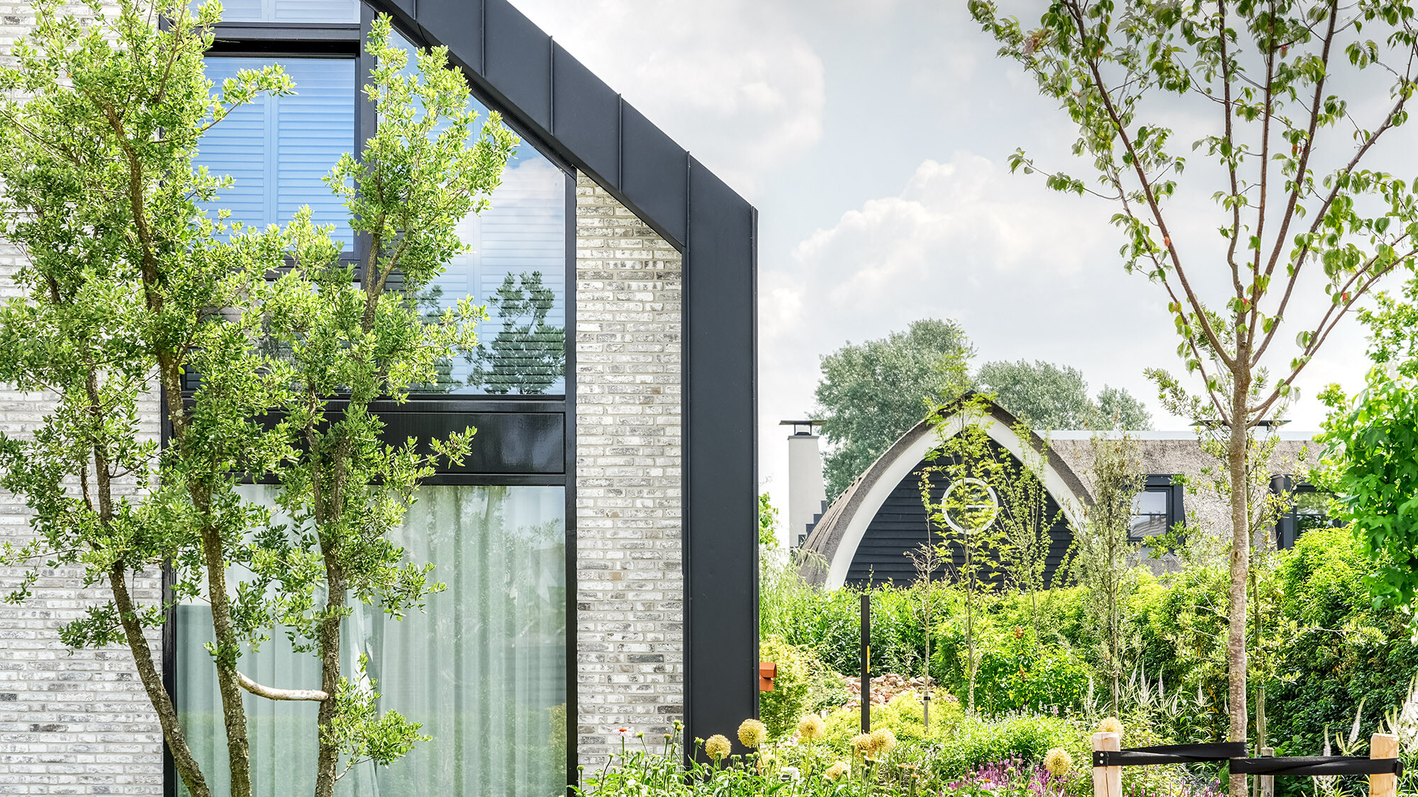 Ein modernes Einfamilienhaus mit schwarzem Prefalz-Dach, eingebettet in einen grünen Garten. Die Fassade kombiniert helle Backsteine mit großen Fensterflächen und dunklen Rahmen. Im Hintergrund ist ein weiteres Haus mit einem ähnlichen Dachstil sichtbar. Der Garten im Vordergrund ist reich an verschiedensten Pflanzen und Bäumen, die dem Bild eine lebendige und naturnahe Atmosphäre verleihen.