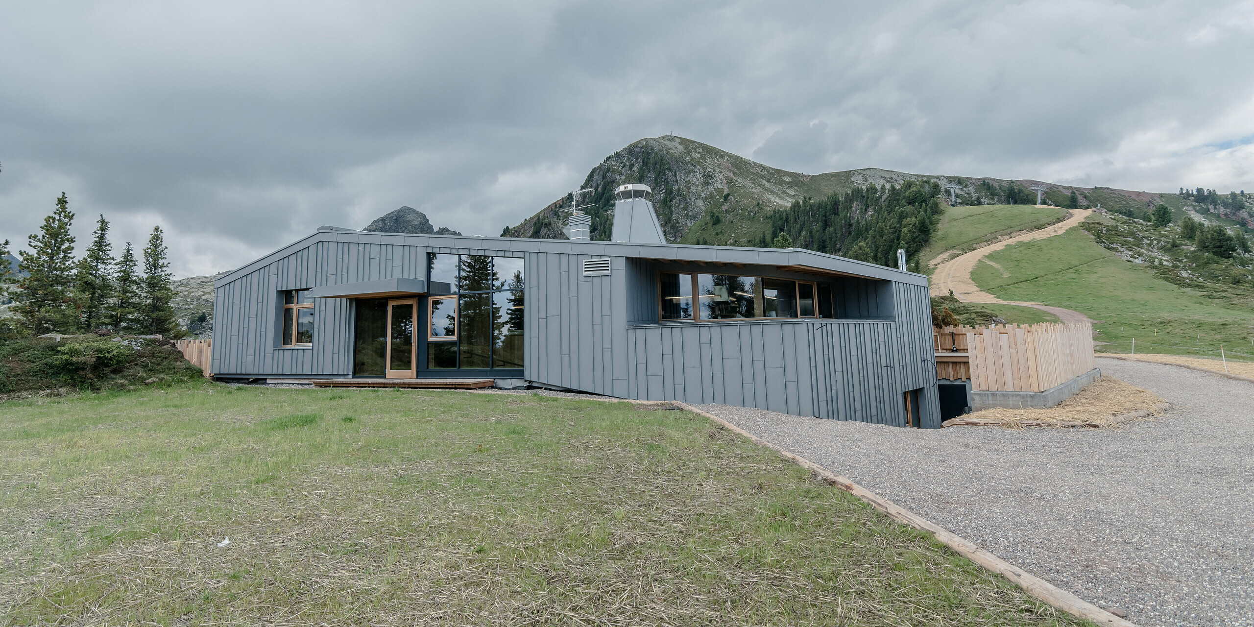 Das Ristorante Busabelle, eingebettet in eine idyllische Berglandschaft, ist von einer Winkelstehfalz Spiegeldeckung aus hellgrauem PREFALZ ummantelt, die die moderne Architektur hervorhebt und eine harmonische Verbindung zur natürlichen Umgebung schafft. Der Eingangsbereich ist mit großen Glasflächen versehen. Im Hintergrund sind Masten für eine Sesselliftanlage zu erkennen. 