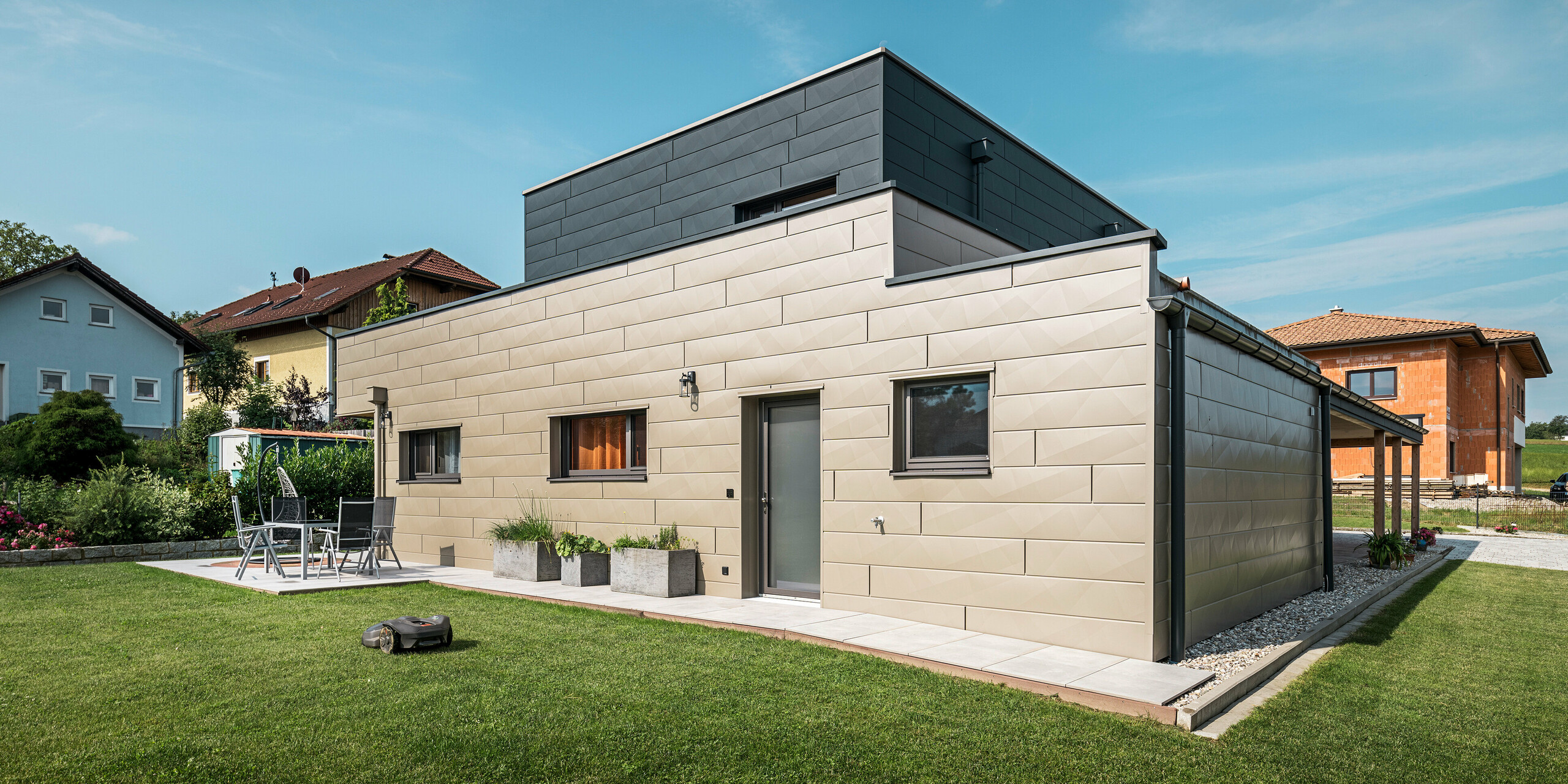 Maison individuelle moderne à Wippenham, en Autriche, avec façade en aluminium PREFA bicolore bronze et anthracite. La façade est conçue avec des panneaux de façade Siding.X, qui assurent un look élégant et contemporain. Le système de gouttière PREFA intégré avec collecteur d'eau et tuyau d'évacuation ainsi qu'un jardin bien entretenu avec terrasse soulignent le design moderne de la maison.