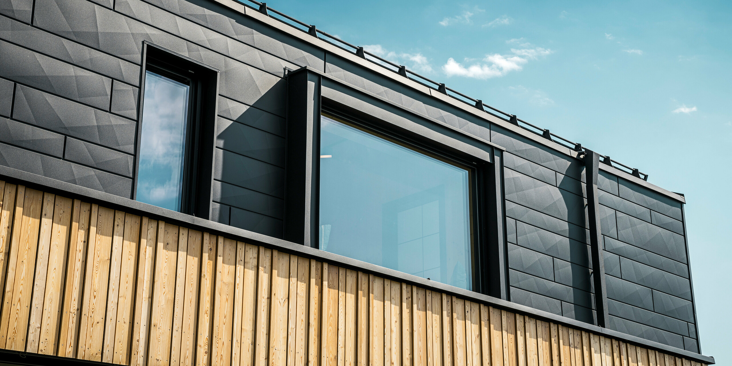 Gros plan de l'étage supérieur de la maison témoin Autark dans le centre de maisons préfabriquées Blaue Lagune, Vösendorf (AT). La façade est constituée de panneaux en aluminium PREFA Siding.X en P.10 noir, qui se distinguent par leur aspect particulier avec des bords différents. En dessous, un élégant revêtement en bois forme un contraste harmonieux avec l'aluminium foncé. Les grandes fenêtres laissent entrer beaucoup de lumière à l'intérieur du bâtiment et soulignent le design moderne.