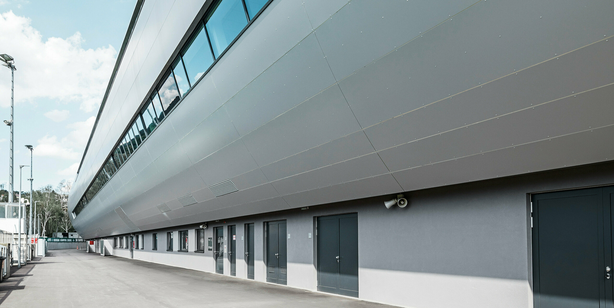 Seitenansicht des Allianz Stadions von SK Rapid Wien, verkleidet mit PREFA Aluminium Verbundplatten in Silbermetallic. Die Fassade zeigt die beeindruckende Länge und klare Linienführung des Bauwerks, während die großflächigen Fenster den Blick zum Trainingsplatz rahmen. In den Fensterscheiben reflektiert der blaue Himmel. Die hochwertige Metallverkleidung verkörpert Beständigkeit und moderne Architektur.