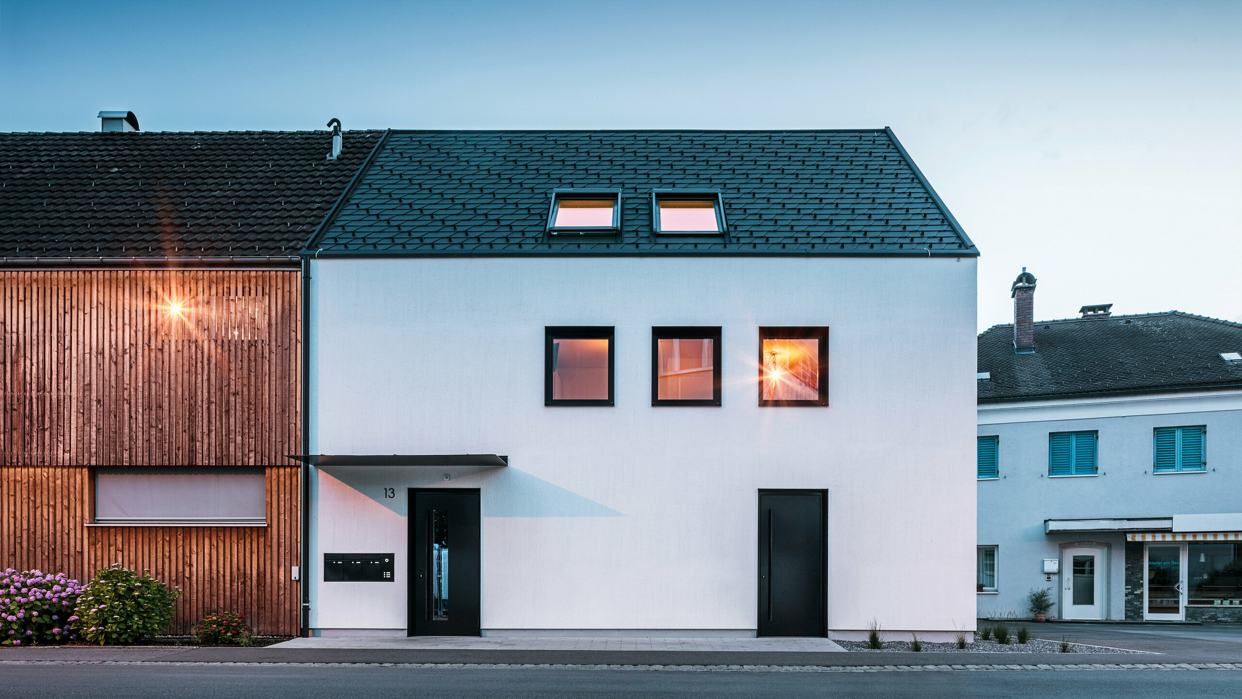 Moderne Mehrfamilienhausfassade mit PREFA Dachschindeln DS19 und Prefalz in Schwarz am Bodensee in Österreich. Das Gebäude hat eine klare Zweiteilung mit einem hölzernen Anbau auf der linken Seite und einem verputzten Hauptteil auf der rechten Seite. Das Dach zeigt die charakteristische dunkle Schindeldeckung und es gibt mehrere rechteckige Fenster, die im warmen Abendlicht spiegeln.