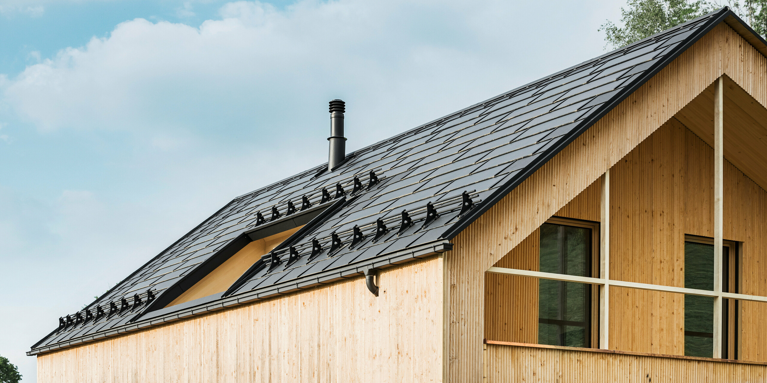 Gros plan sur le pignon d'une maison écologique à Egg, caractérisée par des tuiles solaires PREFA d'un noir élégant, qui s'intègrent harmonieusement au milieu des R.16 PREFA classiques. Les lignes épurées du toit contrastent avec la texture naturelle de la façade en bois, tandis que les tuiles solaires témoignent d'un mode de vie durable. La cheminée subtile s'intègre dans l'ensemble, qui allie un design respectueux de l'environnement et une esthétique rurale. La maison écologique innovante dispose également de deux balcons offrant une vue sur le magnifique paysage montagneux d'Egg dans le Vorarlberg.