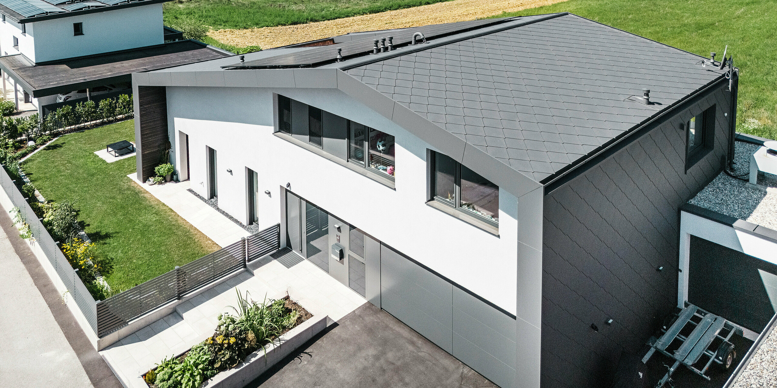 Vue aérienne d'une maison familiale moderne à Aschach avec une architecture intemporelle. La façade et la toiture sont recouvertes de losanges de toiture et de façade 44x44 gris sombre. La coque durable et résistante aux intempéries confère au bâtiment une apparence élégante et offre une protection optimale contre les influences environnementales. Devant la maison se trouve un jardin de devant bien entretenu avec une pelouse verte luxuriante. A droite de la maison il y a une petite dépendance et à côté de la porte d'entrée il y a un garage spacieux avec une allée asphaltée.