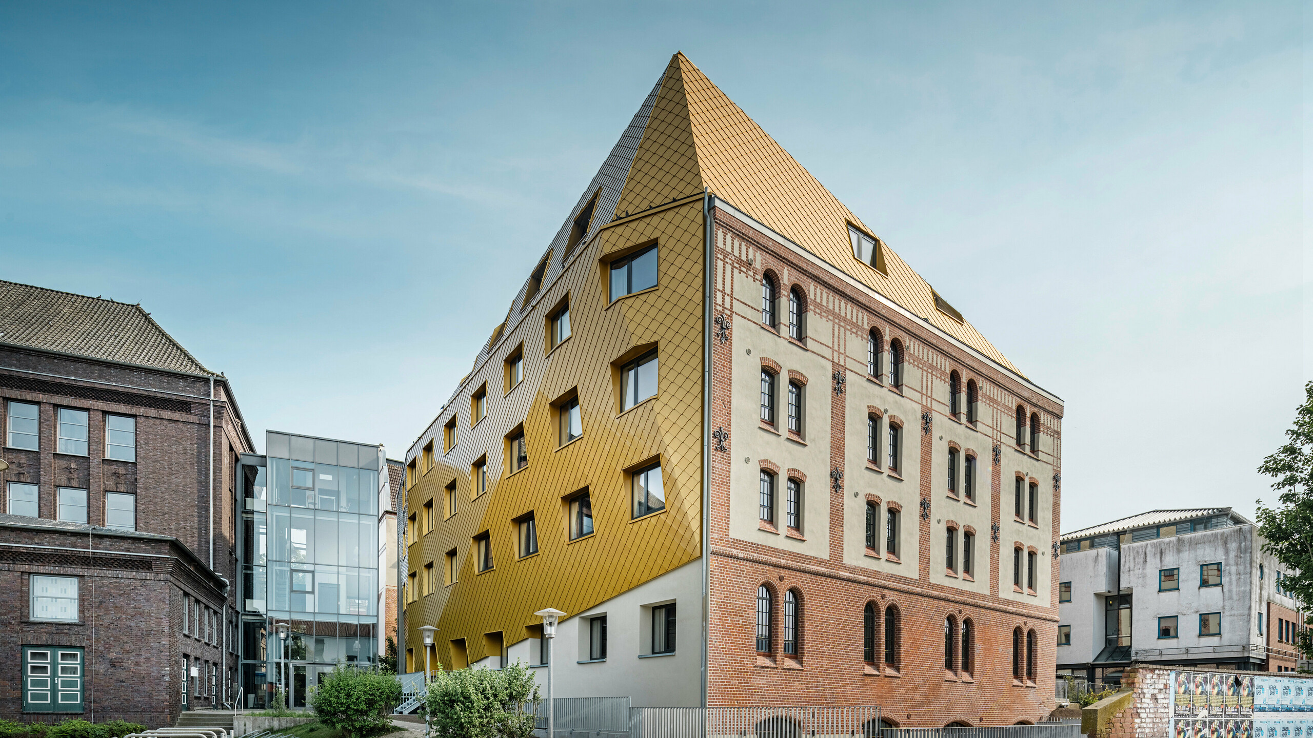 Grondopname van "Het Station" in Kiel. Het studentenhuis is schuin van opzij te zien. De foto laat duidelijk het opvallende contrast zien tussen de aluminium gevel van PREFA dak- en gevellosanges 29 × 29 in Mayagold aan de linkerkant en de onder monumentenzorg vallende pleister- en bakstenen gevel aan de rechterkant.