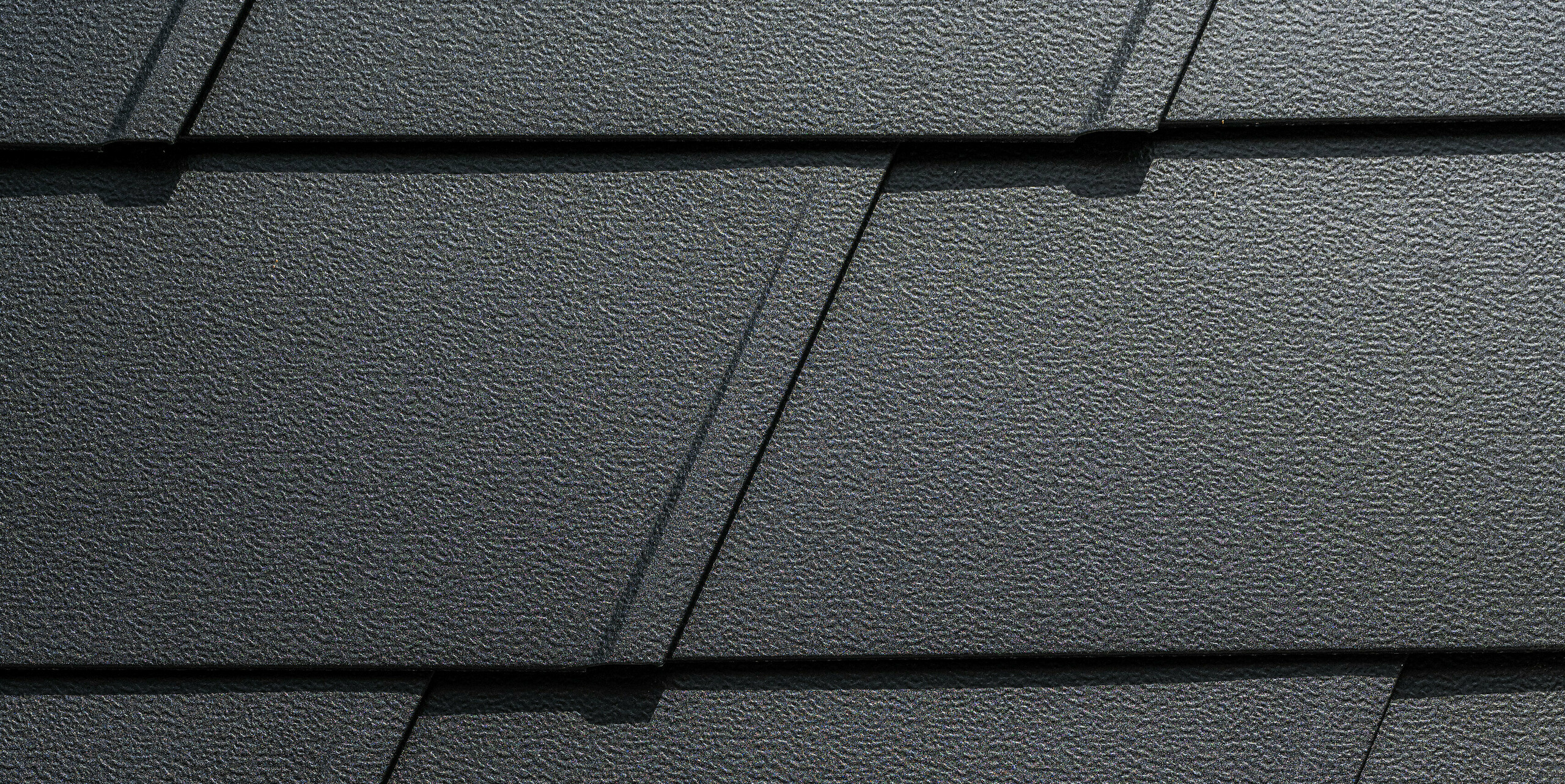 Detailaufnahme eines PREFA Fassadensystems mit schwarzen, strukturierten Aluminiumplatten. Die Wandschindeln sind in einem gleichmäßigen Muster angeordnet, das durch präzise Linien und Fugen betont wird. Dies verleiht der Oberfläche ein modernes und elegantes Erscheinungsbild, das sowohl funktional als auch ästhetisch ansprechend ist.
