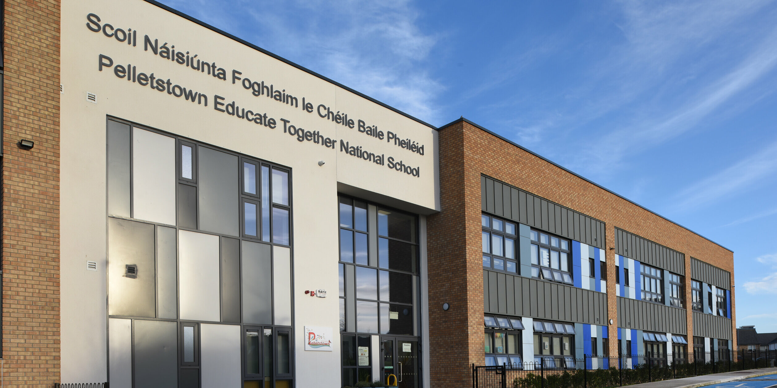 Zone d'entrée de l'école nationale Pelletstown Educate Together à Dublin. La façade de l'école se compose d'un mélange passionnant de matériaux : aluminium, crépi et briques traditionnelles. La toiture à joint debout en PREFALZ en P.10 gris souris souligne l'architecture moderne et durable. Les vélos devant l’entrée montrent que l’école attache une grande importance à la durabilité. Sous les rangées de fenêtres, des arbustes offrent une pause visuelle avec les matériaux de construction robustes.