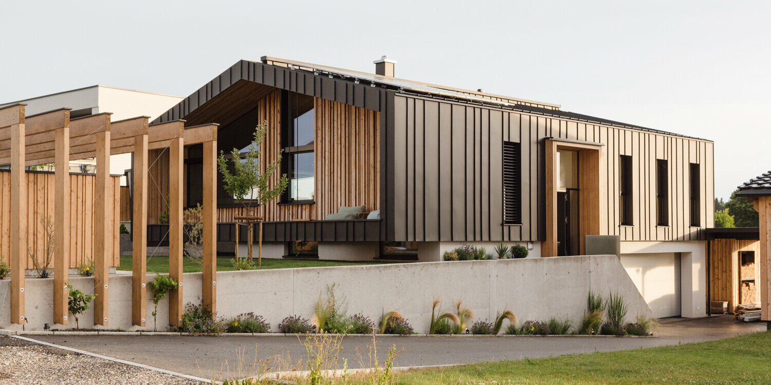 Elegantes Einfamilienhaus in Kirchham, Österreich, mit einer Fassade aus PREFALZ Aluminium in P.10 Anthrazit und strukturierten Holzelementen, die eine warme Atmosphäre erzeugen. Die klare Architektur wird durch die Betonzufahrt und die organisch angelegten Grünflächen ergänzt, welche die moderne Baukunst mit der Natur verbinden. Die präzise Verarbeitung der PREFA Produkte unterstreicht das Engagement für Qualität und Design, was sich in der ansprechenden Ästhetik des Hauses widerspiegelt.